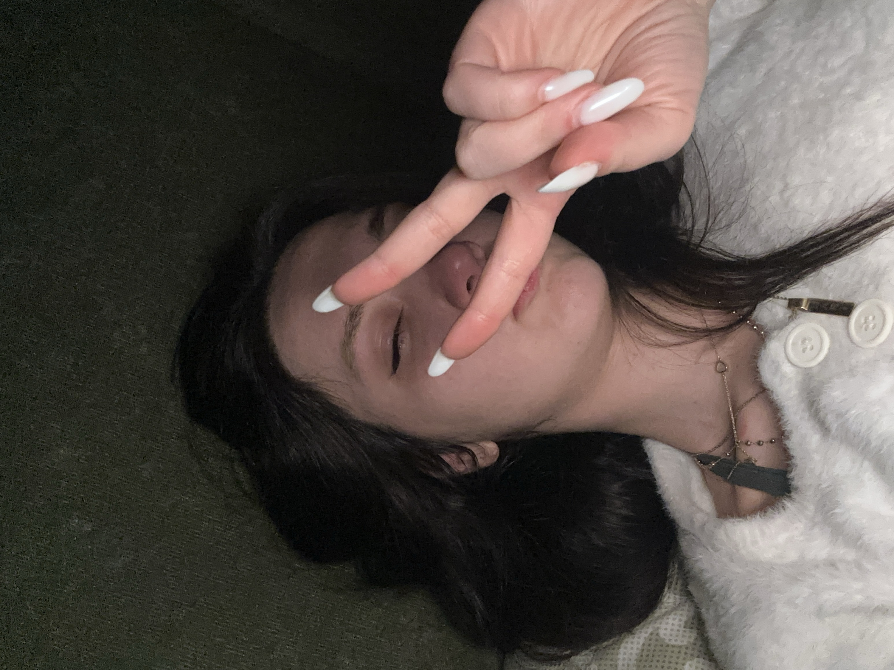 A woman with dark hair lies on a dark green surface. She wears a white fuzzy sweater with white buttons and gold necklaces. Her face is close to the camera, and her eyes are closed. Her hand is raised in front of her face, making a peace sign with long, white almond-shaped nails. The light is low.