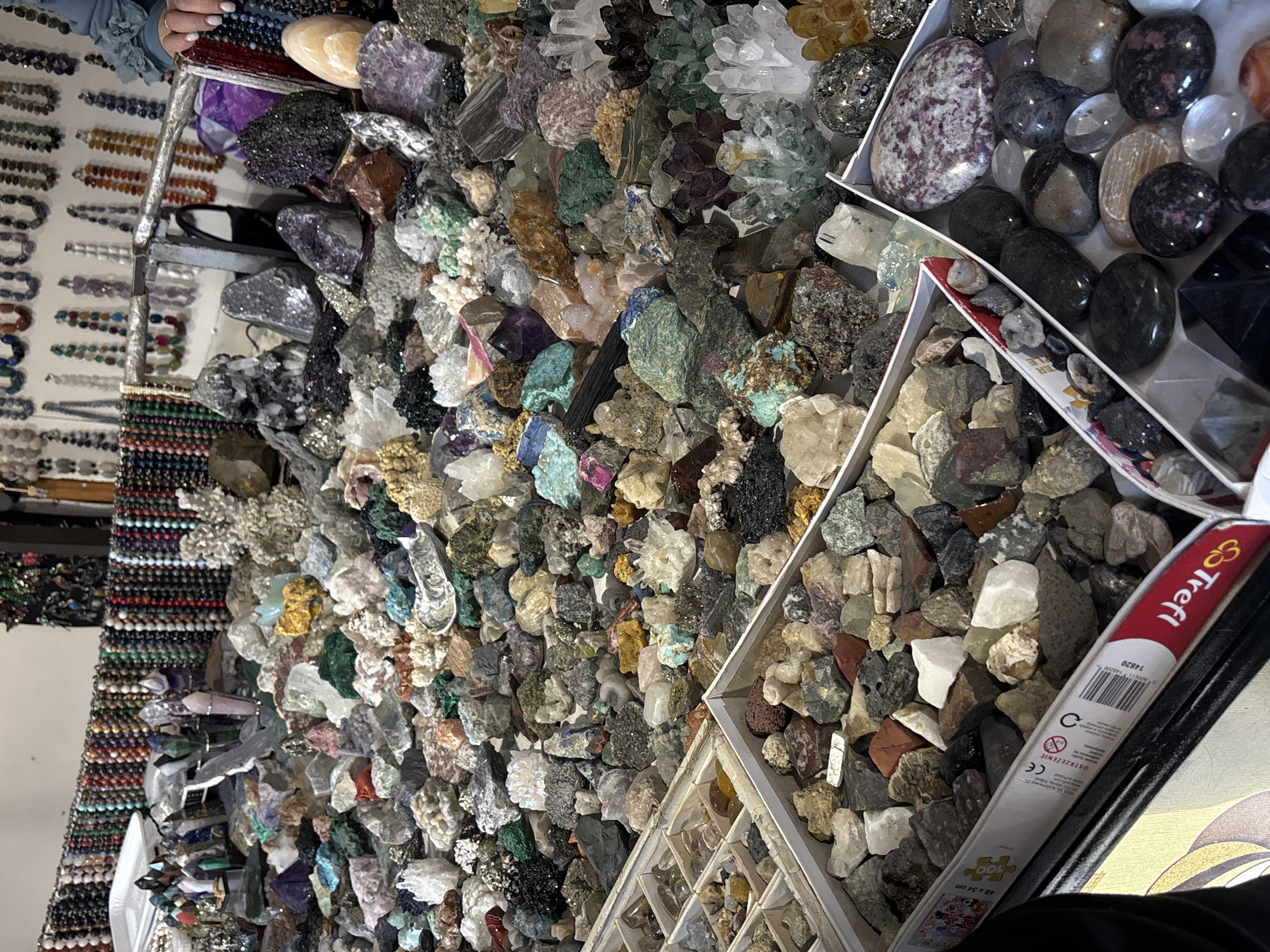 This image shows a market stall in Yerevan, Armenia, displaying a variety of rocks, minerals, and gemstones for sale. The stall is densely packed, showcasing an assortment of items including polished stones, rough crystals, beaded necklaces, and puzzle boxes. The rocks are organized in trays and boxes, suggesting a system of categorization based on type or value. A woman's hand is visible in the upper-left corner, indicating someone is shopping or browsing the merchandise.