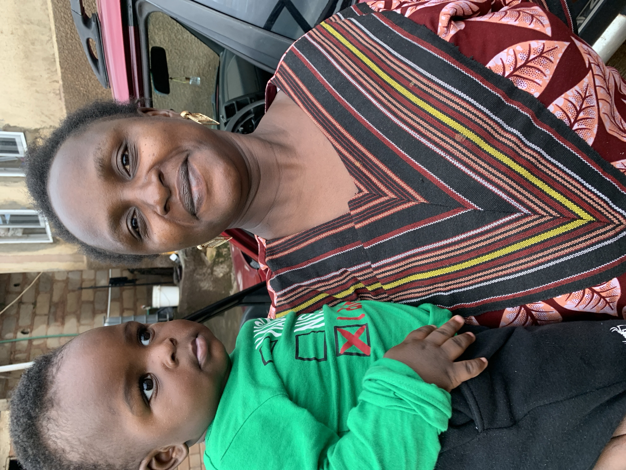 A close-up outdoor medium shot captures an adult woman holding a young child. The woman, who appears to be of African descent, is smiling and looking towards the camera. She has short, dark, curly hair and is wearing large, ornate gold earrings. Her clothing is a patterned fabric with a V-neck, featuring horizontal stripes in black, red, yellow, and white. The child, also appearing to be of African descent, is being held by the woman and is looking to the left of the frame. The child is wearing a bright green long-sleeved shirt with some black and red lettering on the front, and dark pants. Behind them, a red vehicle is partially visible, with its roof rack and side mirror showing. The background consists of a building with visible brickwork and windows, and some green foliage. The lighting suggests it is daytime, possibly in an urban or semi-urban setting. The overall scene conveys a sense of warmth and familial connection.