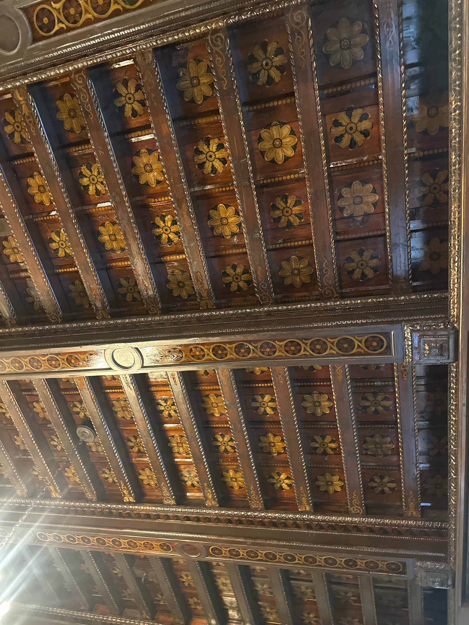 The image shows a close-up of an ornate wooden ceiling, likely from an interior historical building. The ceiling is constructed with dark wooden beams arranged in parallel, with smaller wooden panels placed between them. These panels are decorated with repeating floral motifs painted in yellow and black. The beams themselves are also adorned with gold trim and intricate carvings. A decorative frieze with a pattern of circles and floral elements runs along a horizontal beam in the mid-ground. The overall impression is one of rich, detailed craftsmanship. The location is identified as the Ethnographic Museum in Belgrade, Serbia.