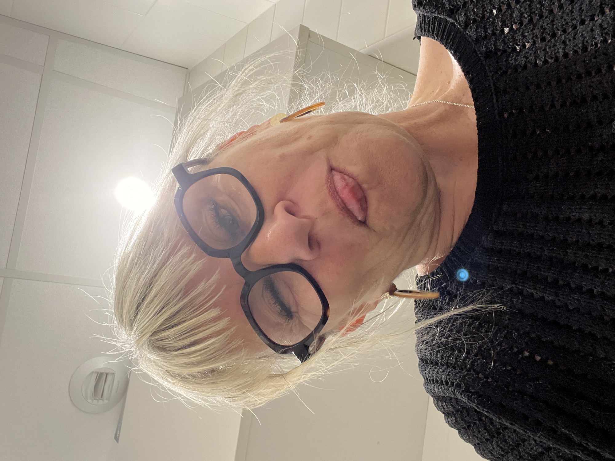 A close-up, downward-angled photograph shows a person with blonde hair and glasses wearing a black, textured sweater. The person's eyes are partially closed, and their tongue is slightly visible. The background includes a white ceiling with a circular ventilation vent. The location is La Fouillouse, France. No specific actions or interactions are evident in the photograph.