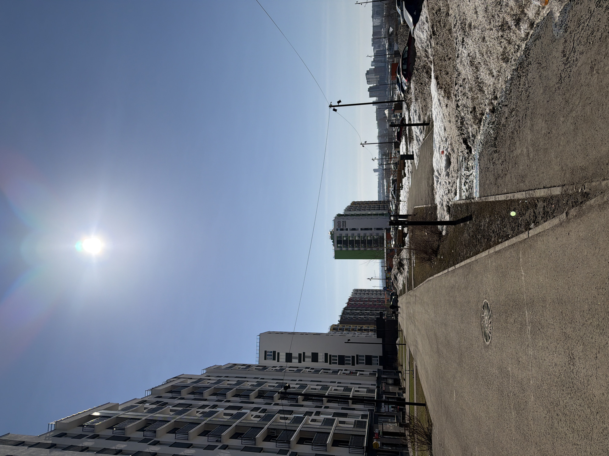 The image shows an outdoor urban scene in Pervomaysky, Russia, on a bright, sunny day. A gray asphalt pathway runs along the right side of the frame, displaying a circular manhole cover near the center. Adjacent to the pathway is an area covered in melting snow, patches of bare earth, and low vegetation.
The left side of the image features contemporary high-rise apartment buildings with white facades and dark balconies. In the distance, other apartment buildings of varying heights and colors, including one with a green accent, are visible. Utility poles with visible wires run along the scene, slightly offset from the building line. The clear, blue sky occupies the upper portion of the image, with the sun appearing as a bright, slightly diffused circle on the left. No people are clearly visible in the image.
