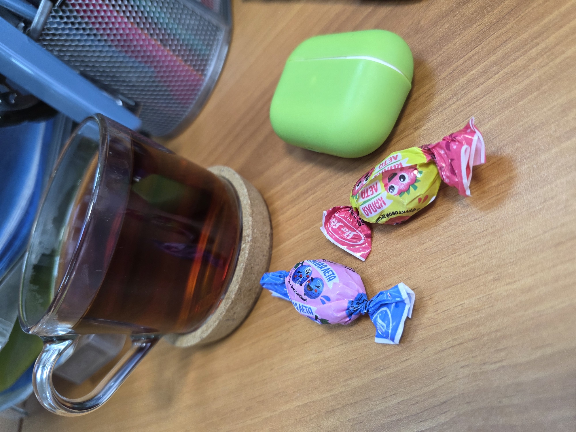 The image captures a close-up, eye-level view of several items arranged on a light-brown wooden desk surface in Chaikovsky, Russia. In the foreground, to the left, a clear glass mug filled with dark brown tea rests on a round cork coaster. The tea shows reflections of ambient light.
To the upper right of the mug, a bright lime-green wireless earbud case, likely for AirPods, sits closed. Below and to the right of the earbud case are two individually wrapped candies. The top candy is wrapped in yellow and pink foil, featuring a cartoon strawberry or raspberry design. Partially visible text on this wrapper reads "ЯГОДНОЕ ЛЕТО" (Berry Summer) and "ФРУКТОВЫЙ" (Fruity), along with the brand name "КРАСНЫЙ ОКТЯБРЬ" (Red October). The second candy, positioned below the first, is wrapped in pink and blue foil, displaying cartoon blueberries. Its wrapper also shows text, including "ЯГОДНАЯ ЛЕТО" (Berry Summer) and "ФРУКТОВЫЙ НАЧИНКОЙ" (Fruity Filling).
In the upper left background, part of a gray metallic mesh office organizer is visible, holding colorful pens or pencils. A blue plastic or metal arm of another office item runs diagonally behind the mug and in front of the organizer. The lighting is soft and even, suggesting an indoor setting during the day. The scene depicts a casual break, possibly from work or study, with tea and sweets at hand.