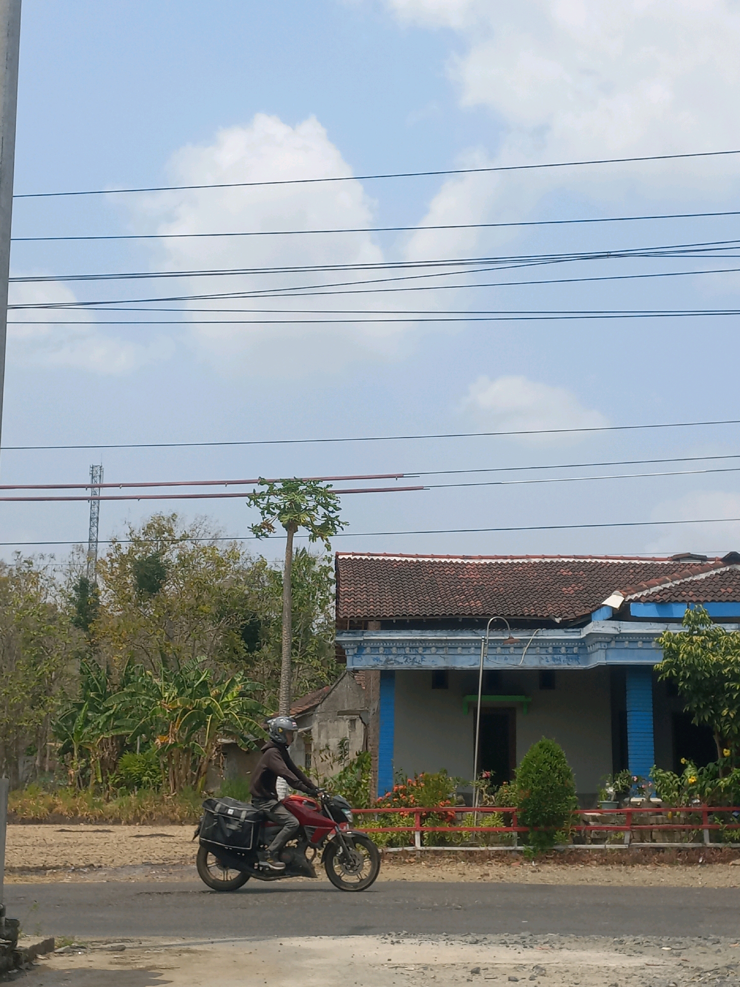 A person wearing a helmet rides a red motorcycle with two black saddlebags in the city of Baturetno, Indonesia. The person is wearing a dark brown jacket and grey pants. The motorcycle is on a paved road, which appears to be wet in some areas. In the background, there is a blue and white house with a red tile roof. There are also trees and power lines visible in the scene. The sky is blue with some white clouds.