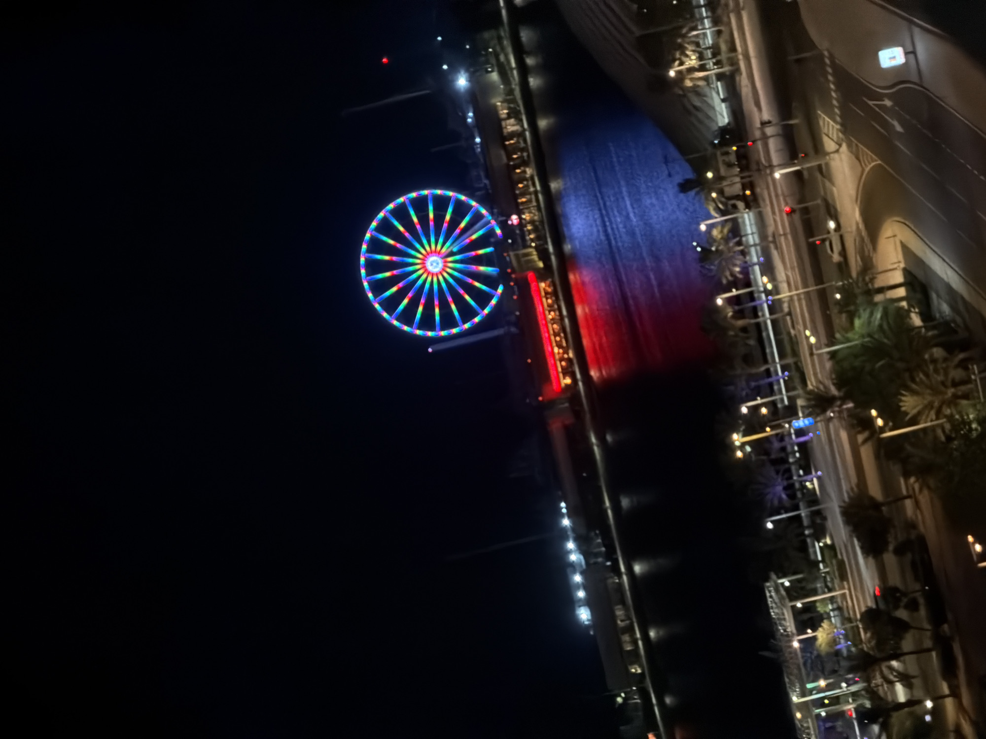 In the city of Unknown, Unknown, at night, a Ferris wheel illuminated with multicolored lights dominates the scene. The Ferris wheel is situated near a body of water, reflecting the vibrant lights of the surrounding structures. Buildings with illuminated windows and red architectural lights are visible along the waterfront. Palm trees and streetlights line a paved area with roads and sidewalks in the foreground, suggesting an urban entertainment district. Vehicles are present on the roads. The sky is dark, indicating it is nighttime.