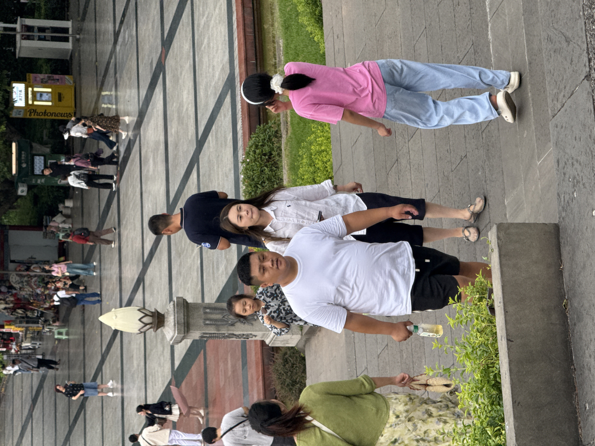 The image shows a group of people walking on a paved plaza in Yuzhong District, China. In the foreground, a man in a white t-shirt and black shorts is walking towards the right, holding a bottle of yellow liquid. To his left, a woman in a green top and a floral skirt is walking away from the camera, carrying a woven bag. Behind them, a young girl in a patterned dress is looking down. Further back, a woman in a white collared shirt is walking to the left. A woman in a pink t-shirt and blue pants is walking away from the camera on the right. In the background, other individuals are scattered across the plaza, some carrying bags and wearing casual clothing. The plaza is paved with large rectangular tiles and has dark lines separating them. A lamppost with a lotus-shaped top is visible in the middle ground. Green bushes and a body of water are present to the right of the paved area. The overall setting appears to be a public outdoor space in a city.