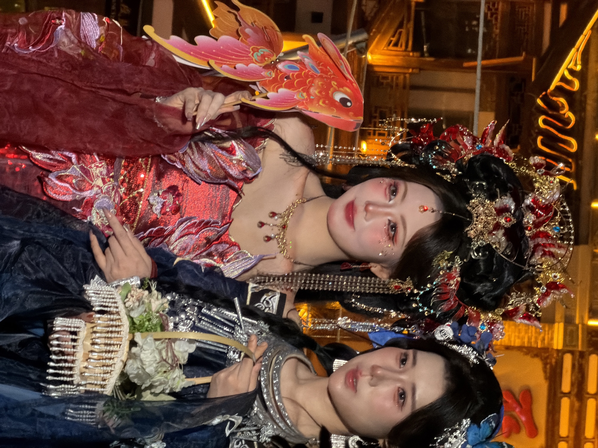 Two young East Asian women are posing for a photograph in front of traditional Chinese architecture illuminated by warm lighting. The woman on the left wears an elaborate red and gold headdress with dangling jewels and chains, a red sequined top with ruffled sleeves, and a sheer red overlay. She is holding a decorative red and orange paper koi fish. Her makeup is dramatic with glitter accents on her eyes and cheeks. The woman on the right has a blue butterfly hair ornament and is wearing a dark blue garment with silver embroidery and trim around the neckline and sleeves. She is holding a small woven basket filled with white and green artificial flowers. Both women have their hair styled in dark updos. The background shows intricately decorated wooden eaves and structures, suggestive of a historical district in Yuzhong District, China. The overall atmosphere appears festive, likely during a cultural event or celebration.