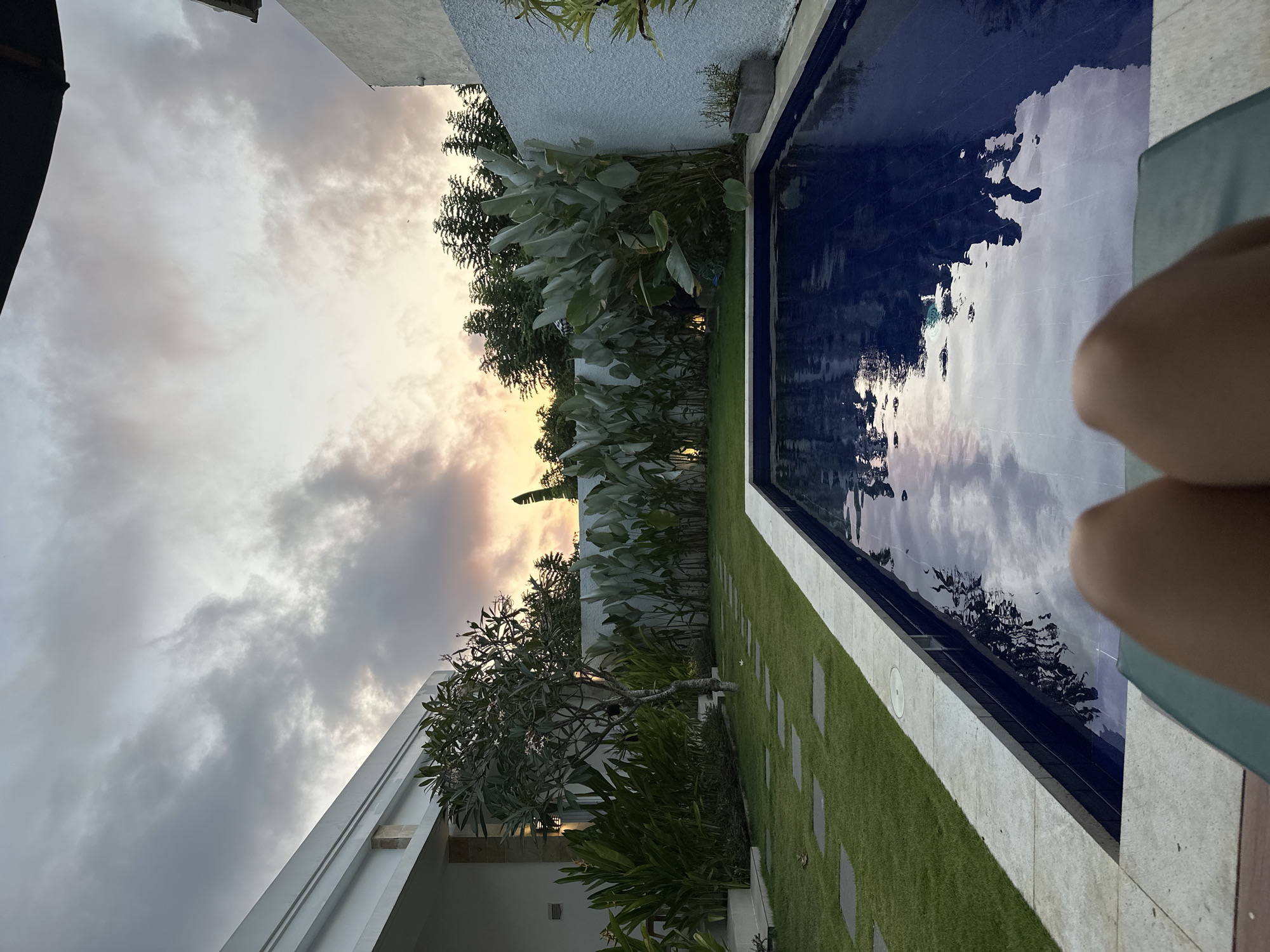 In the city of Kuta Selatan, Indonesia, a person relaxes on a lounge chair facing a private swimming pool as the sun sets. The pool's dark blue water reflects the cloudy sky and surrounding greenery. A well-maintained lawn with stepping stones leads to the pool, bordered by a white tiled edge. Lush green plants and a small tree are situated in the yard, with a modern white building visible on the left. A textured white wall encloses the backyard, with a planter holding small green plants on the right. The sky is filled with dramatic clouds tinged with the warm colors of sunset.