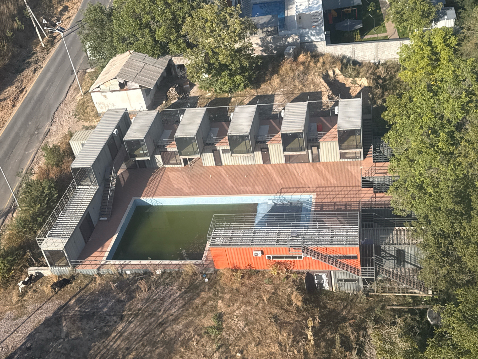 The aerial photograph shows a rural or semi-rural setting with a mix of structures, vegetation, and a road. In the foreground, two cows are grazing on dry, brown vegetation. To the right of the cows, a large swimming pool with green water is visible, bordered by a tiled deck and a metal railing. Adjacent to the pool is an orange structure with an air conditioning unit and a window, likely a container-style building. Behind the pool area, a complex of modular, metal-clad units, possibly shipping containers converted into rooms or studios, are arranged in a row. These units have glass windows and metal stairs leading to them. Further back and to the left, a dilapidated building with a damaged roof sits near a paved road. The road curves and appears to be in disrepair in some sections. Lush green trees are scattered throughout the scene, and the ground is covered in a mixture of dry grasses, dirt, and some rocks. In the upper right corner, another structure, possibly a multi-story building with a courtyard or garden area, can be seen. The overall impression is one of a developing or rustic commercial establishment, possibly a resort or accommodation, situated on the outskirts of Amirsay, Uzbekistan.