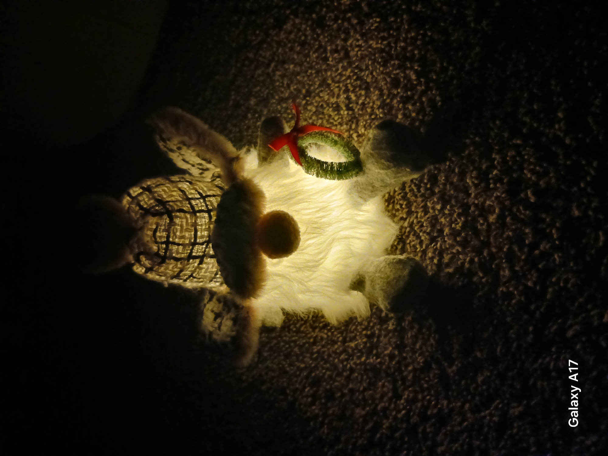The image shows a stuffed animal lying on a textured surface, likely a rug or carpet, in a dimly lit environment. The toy features a patterned body, possibly of a plaid design. It has fuzzy, light-colored limbs and a spherical, yellow pom-pom. A small, green and red decorative ring rests near the head of the animal, perhaps indicating a festive theme. The light source appears to be emanating from underneath the toy. The words "Galaxy A17" are visible in the lower right corner of the image, indicating the model of the camera used to take the picture. The location is unknown, not identifiable as Yekaterinburg, Russia, and no people, actions, or interactions are evident.
