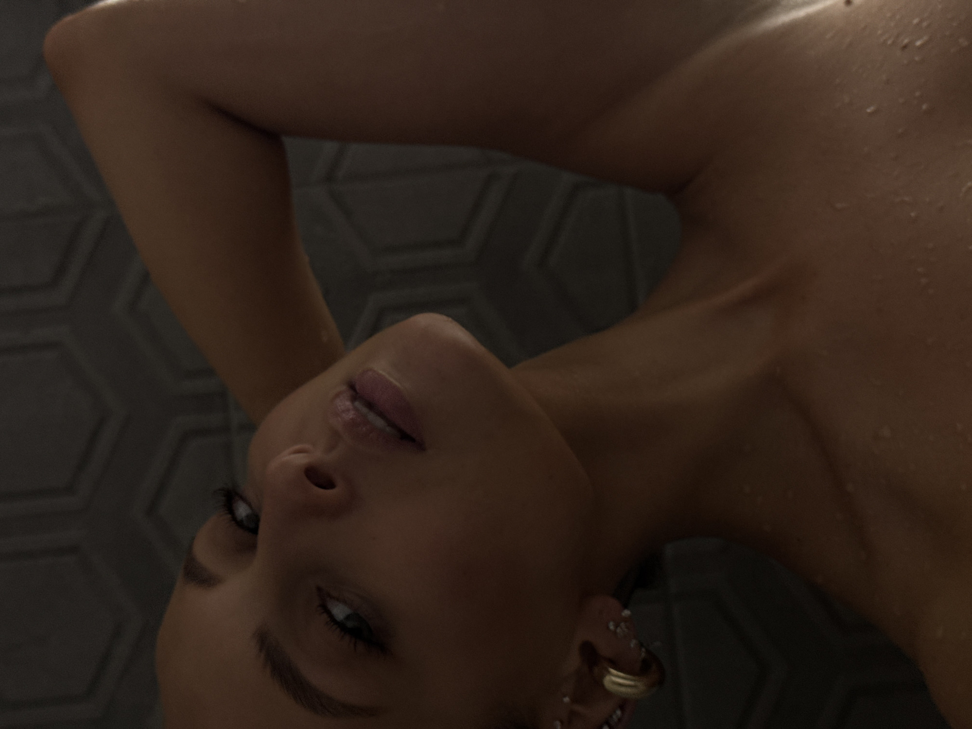 The image is a close-up, low-angle shot of a woman's face and upper body. She is looking upwards and to her left, with her right arm raised behind her head. Her skin appears wet, suggesting she may be in a shower or has recently been in water. She is wearing hoop earrings and several smaller stud earrings in her left ear. The background consists of a textured wall, possibly tile, with a hexagonal pattern. The lighting is dim and dramatic, with the woman's face partially illuminated, creating shadows. The image does not contain any information about the city of Chaikovsky, Russia.