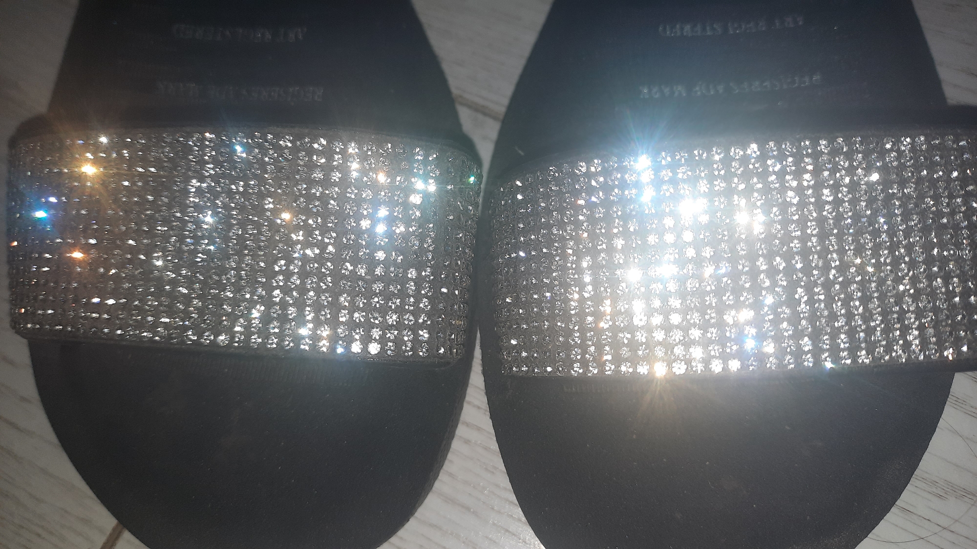 This image shows a close-up, top-down view of two black slide sandals with rhinestone-encrusted straps. The sandals are placed on a light-colored wooden surface, possibly a floor. The rhinestones on the straps are highly reflective, causing numerous bright glints and lens flares throughout the image, particularly on the upper sandal. The lighting appears to be direct and bright, creating strong highlights and shadows. Text is visible on the inside edge of the straps of both sandals, but it is largely obscured by the shine of the rhinestones and the angle of the photograph. The text appears to be embossed or printed in a light color.
Without further information, it is impossible to determine the location within the city of Шклоў, Belarus. The image focuses solely on the footwear and its immediate surface.