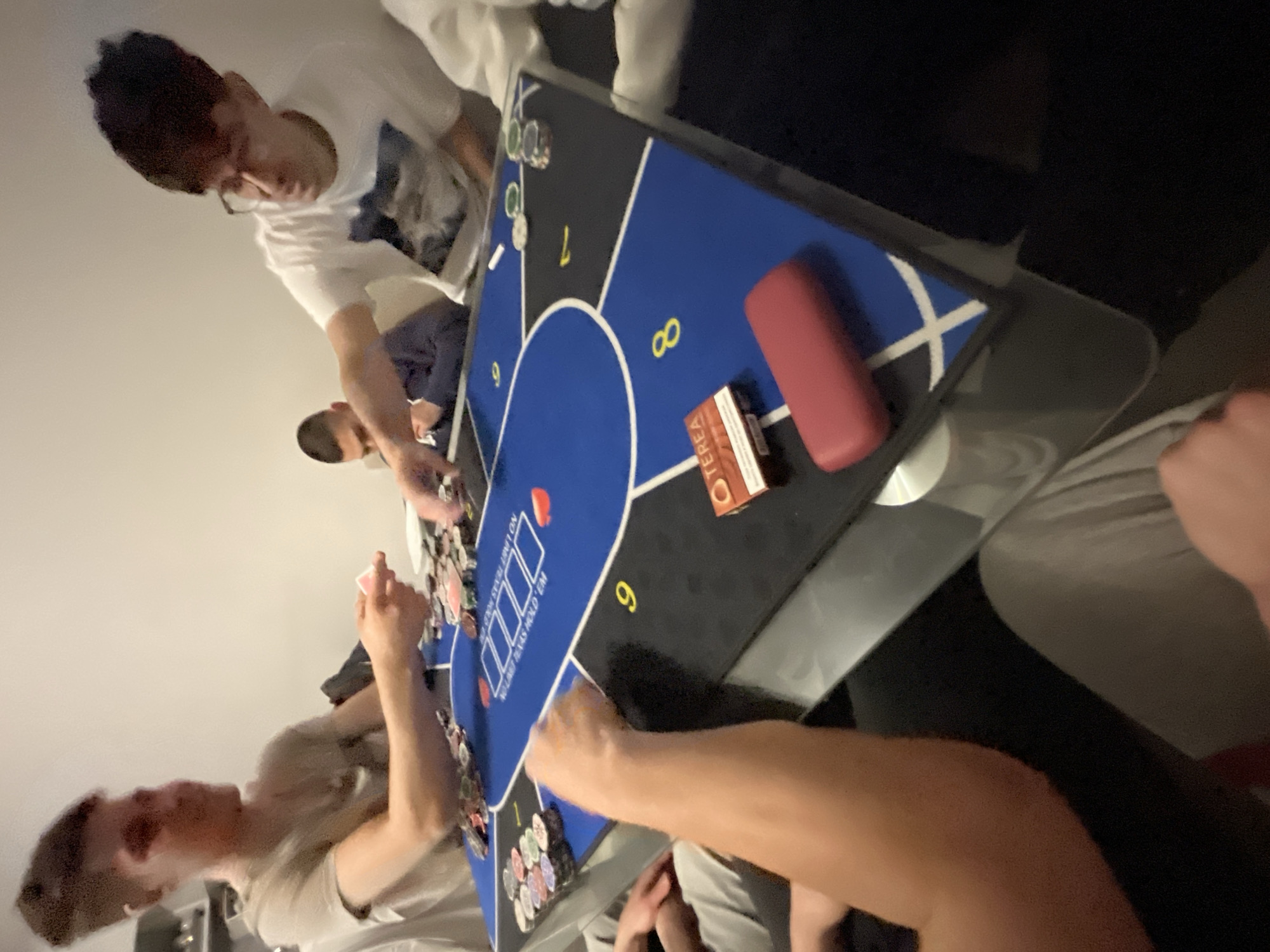 Several people are playing poker at a table. There are poker chips and cards on the table. A pack of cigarettes and a lighter are also on the table. The location is unknown.