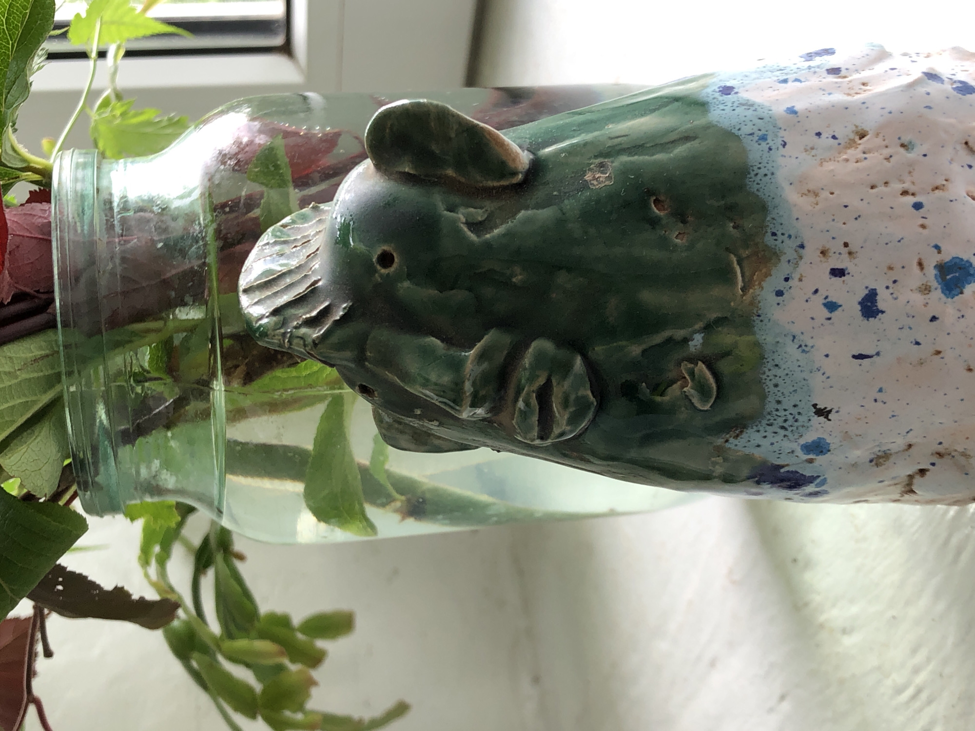 The image displays a unique indoor plant propagation setup, likely placed on a windowsill and illuminated by natural daylight. A clear glass bottle is positioned horizontally, with its opening towards the left, containing water and several green and reddish-dark green plant cuttings.
Attached to the right side of the bottle is a decorative ceramic object shaped like an animal head, possibly a stylized pig or bear. This ceramic piece features distinct ears, a textured snout with small nostril-like holes, and subtle facial contours. The animal head is glazed in a mottled, dark green color, which gradually transitions into a lighter blue edge before becoming a rough, porous white texture. This white section is heavily speckled with various shades of blue dots, creating a cloudy or abstract pattern. The ceramic piece appears to be integrated with or resting on the bottle, serving as both a decorative element and possibly a counterweight.
In the background, a white window frame and a plain white surface, consistent with a windowsill, are visible. The scene is brightly lit, suggesting daytime. There is no visible text in the image.