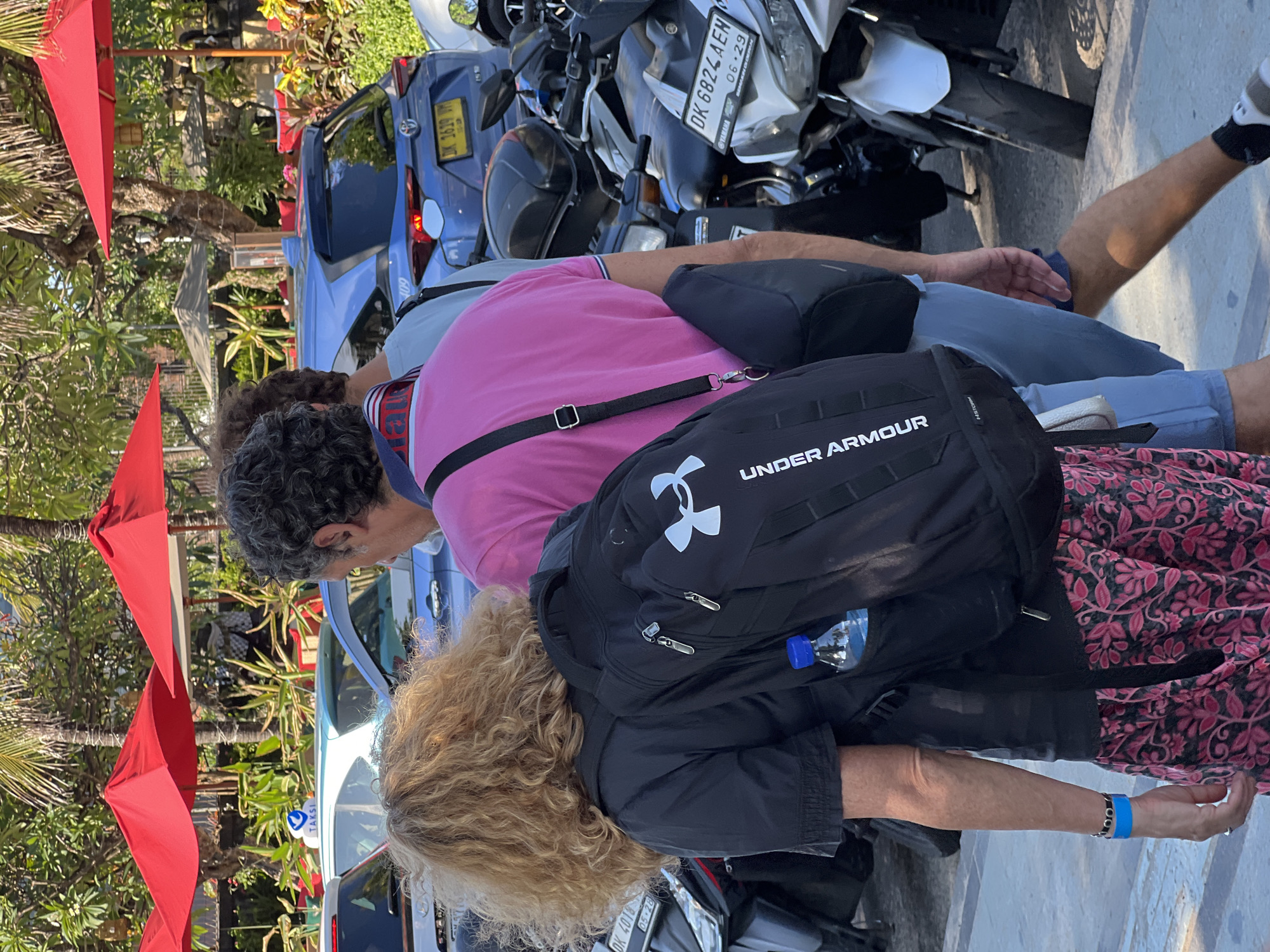 In Kuta, Indonesia, a group of people walk past parked motorcycles and vehicles under red umbrellas. A woman with curly blonde hair, wearing a black shirt and a patterned skirt, carries a black backpack with the Under Armour logo. She has a blue wristband and a bracelet on her left arm. A man in a pink shirt and blue shorts walks beside her, carrying a dark bag across his chest. Another person in a light blue shirt is partially visible behind him. The background includes palm trees, foliage, and several cars, including a blue Toyota with a license plate starting with "DK 3639". The scene appears to be a street or walkway in a busy area.