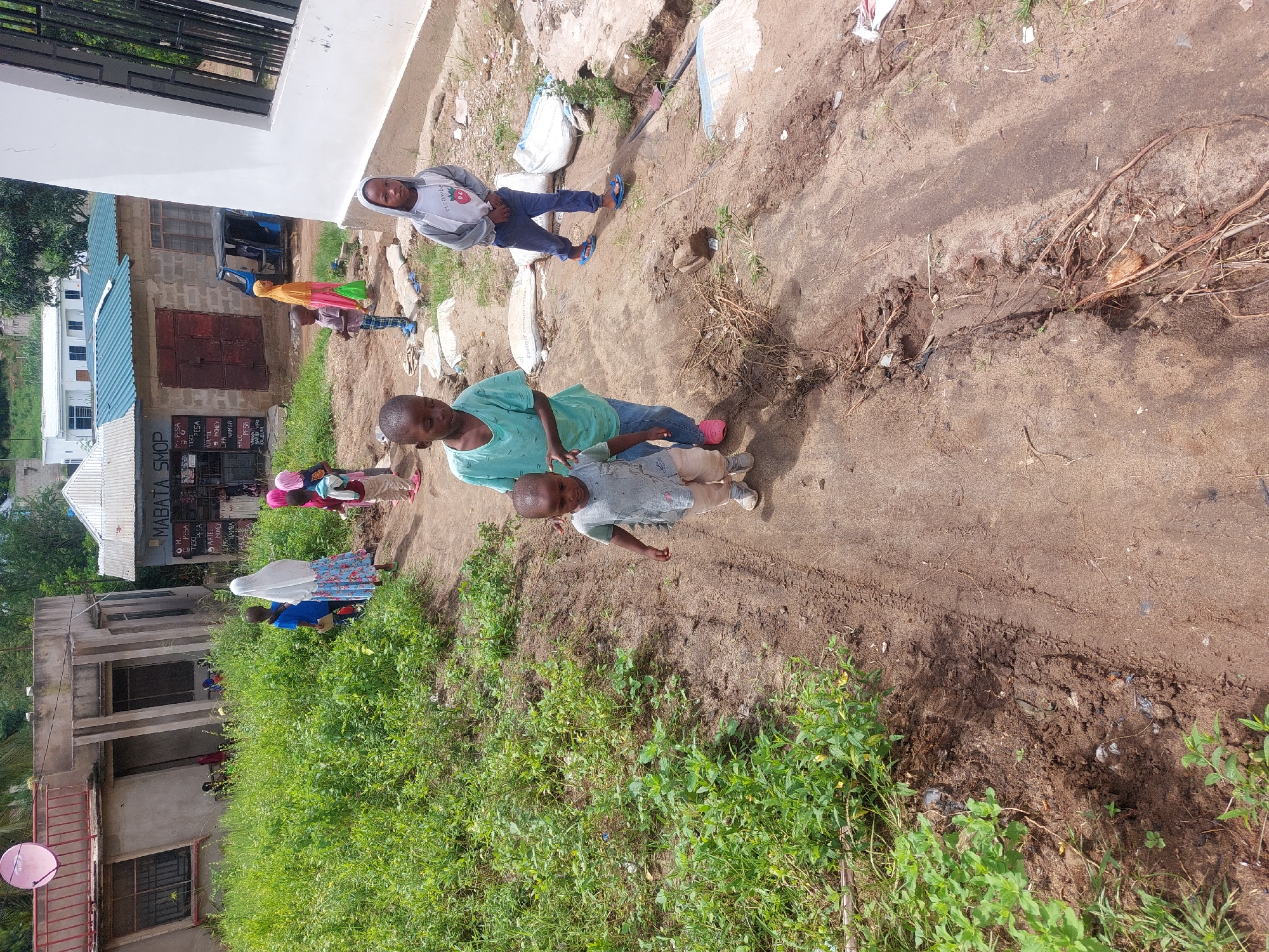 The image shows a dusty path in an urban area, likely in Dar es-Salaam, Tanzania, given the visible signs of Tanzanian commerce such as the "Mabata Shop" sign advertising "Tigo Pesa" and "Airtel Money" (mobile money services). The path is bordered by low-lying vegetation and a building on one side, and by construction materials and a partially constructed or run-down building on the other. Several people are present, including children and adults. A woman and child are in the foreground, while a group of people appear to be walking toward a storefront in the background. Construction materials (likely cement bags) are scattered along the path.