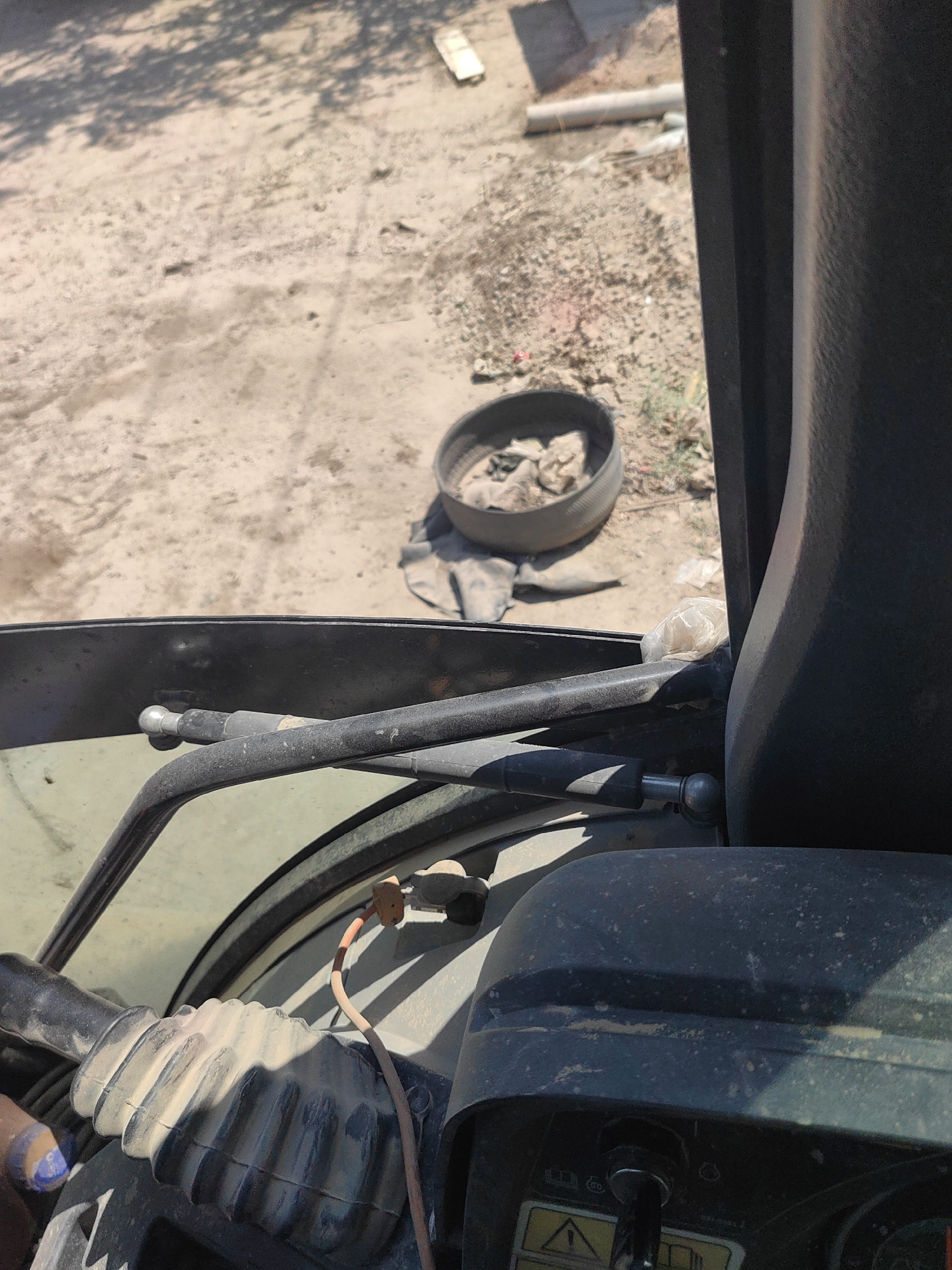 The image shows a close-up view of the interior of a heavy machinery vehicle, likely a tractor or construction equipment, with the exterior visible through the open door or window. The interior is dusty and shows parts of the steering column, levers, and a dashboard with a key in the ignition. A red wire with an orange connector is visible, attached to a component on the dashboard.
Outside the vehicle, the ground appears to be dirt and gravel, with some scattered debris including what looks like an old tire with rags inside, and some construction materials like pipes. The lighting suggests it is daytime with bright sunlight, casting shadows from overhead foliage. The scene appears to be in an outdoor work area, possibly a construction site or a rural property. The city of Shymkent, Kazakhstan, is not directly depicted or identifiable within the provided image content.