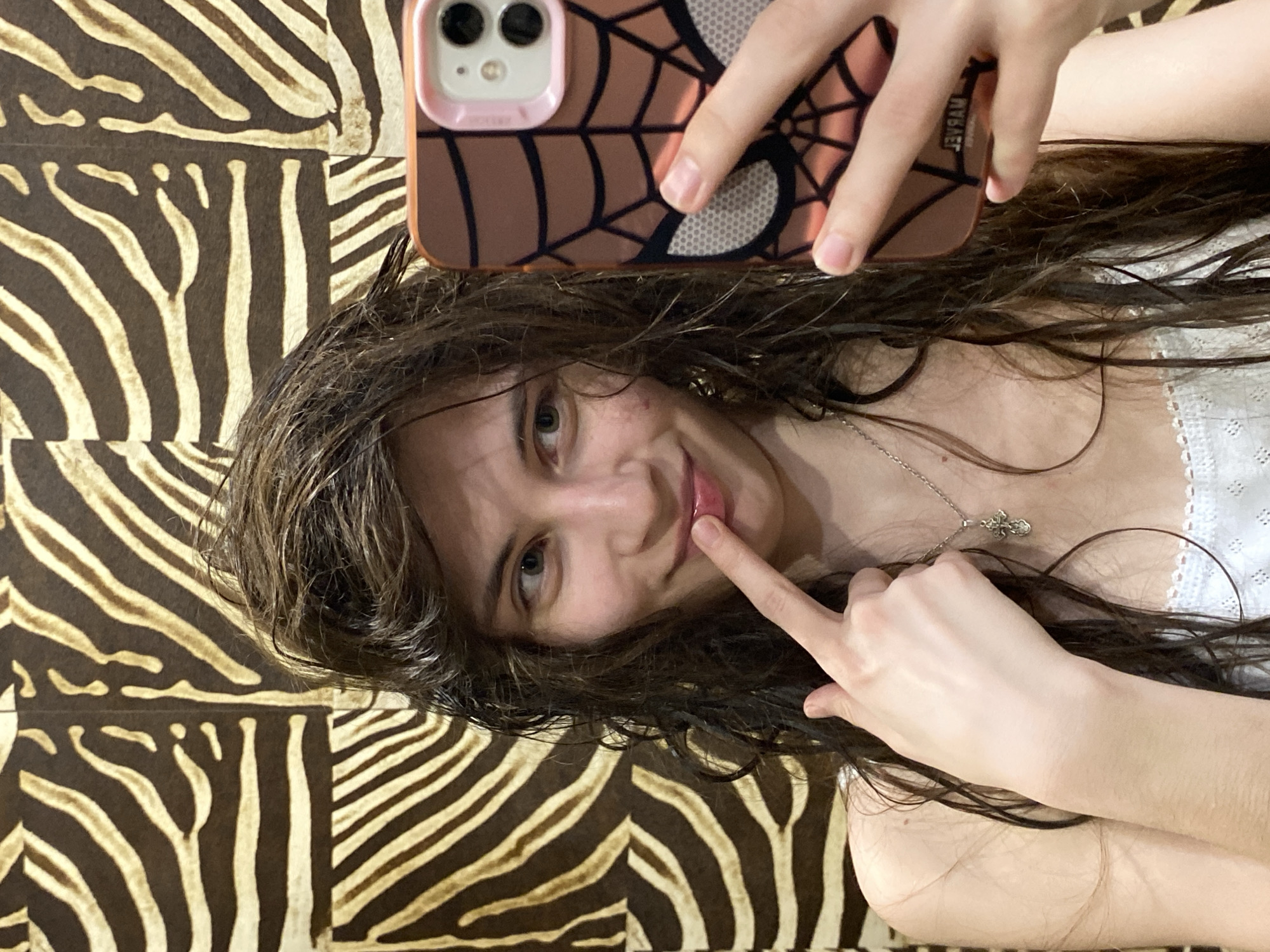 A selfie shows a young woman with long, dark, wavy hair, which appears wet, captured indoors in Votkinsk, Russia. She is wearing a white top with a delicate lace or embroidered trim and a silver necklace with a small cross pendant. The woman holds a smartphone, visible in the upper part of the frame, encased in a distinctive brown case featuring a spiderweb design and a pink trim around its dual camera module. Her right hand grips the phone, while her left index finger gently touches her lips, accompanying a soft smile as she looks towards the camera. The background is composed of a prominent pattern of brown and beige/gold zebra stripes, suggesting a patterned wall or textile. Faint, mirrored text, possibly "NYSAW" or similar, is visible on the phone case but is not clearly legible. The overall impression is a casual, personal moment captured indoors.