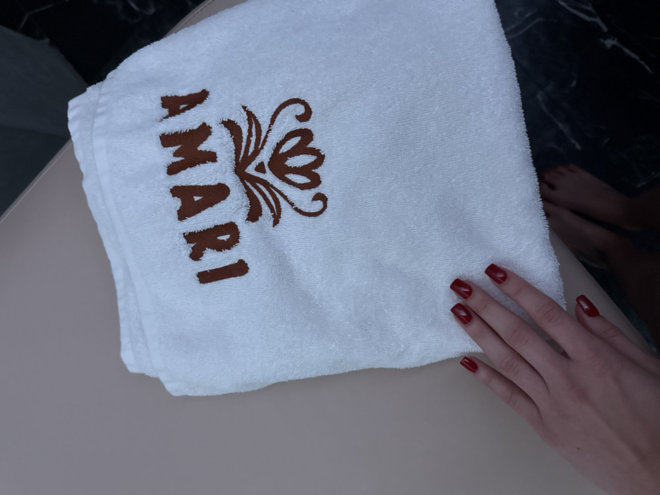 An interior scene in Kyiv, Ukraine, depicts a white, folded terry cloth towel with the word "AMARI" embroidered in brown uppercase letters and an accompanying ornate brown floral design. The towel is situated on a light beige surface. A human hand, featuring red-painted fingernails, is positioned in the lower-left foreground, resting on the towel's edge. In the lower-right, two bare human feet are partially visible on a dark, veined surface, consistent with marble flooring.