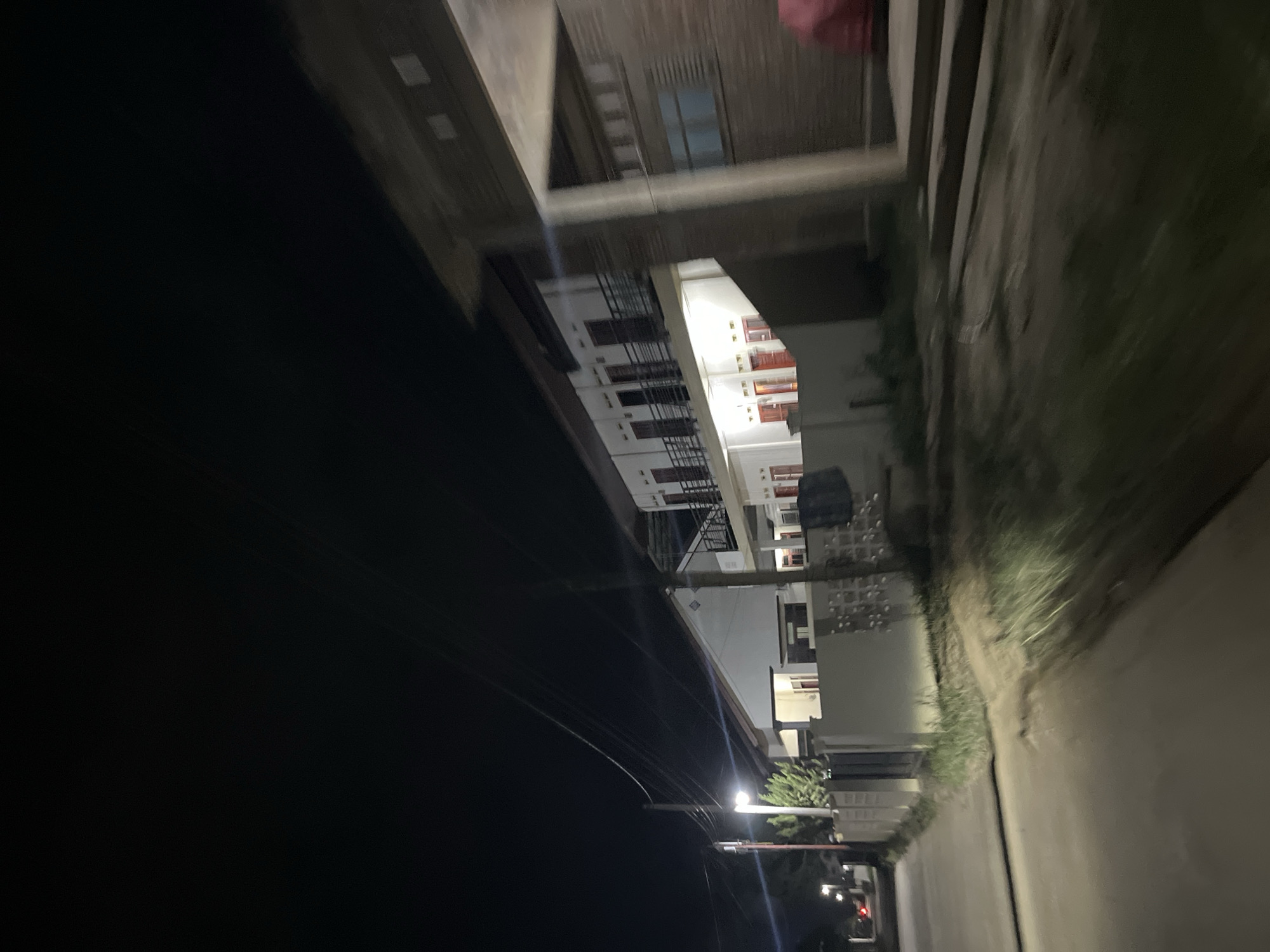 This media file captures a nighttime view from a vehicle moving along a street in Menanggini, Indonesia. The scene is dominated by the dark sky and illuminated buildings.
The primary subject is a multi-story building with a white facade, featuring multiple windows and balconies with metal railings. Some windows are open, revealing glimpses of interiors. Red-colored doors are visible on some levels. The building appears to be an apartment complex or a guesthouse.
In the foreground, power lines are strung across the scene, some illuminated by a street light. The street itself is visible, with a grassy area on the right side of the frame, suggesting the presence of vegetation. The vehicle's movement is indicated by motion blur, especially in the right half of the image, and the overall lighting suggests it's nighttime.
There are no visible people in the image. The weather appears to be clear, as there are no indications of rain or fog. The overall atmosphere is quiet and residential, characteristic of a town at night. No text is visible in the image.