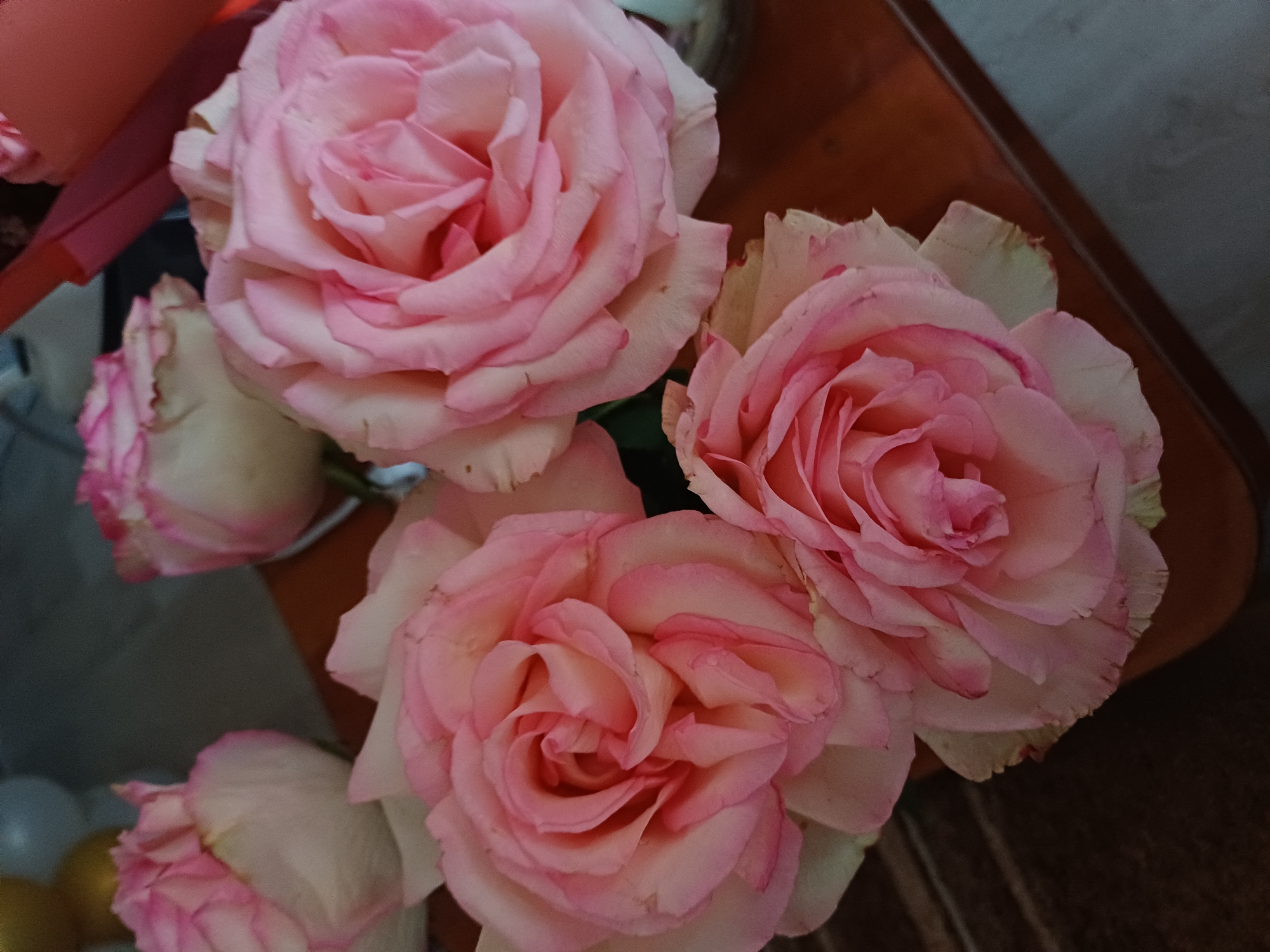 A close-up shot shows a bouquet of pink and white roses. The roses are in various stages of bloom, with some fully open and others still tightly budded. The petals are a soft pink, with white edges and hints of green on the outer layers. The roses are arranged in a way that suggests a gift or a decorative display. The background is a mixture of dark wood and a gray surface, with a hint of a brown object, possibly a table or a vase. The lighting is soft, creating a gentle glow on the petals. There is no text visible in the image, and no people are present. The overall impression is one of natural beauty and elegance. The image appears to be taken indoors, possibly in a home or a floral shop. The focus is entirely on the roses, highlighting their delicate textures and soft colors. The subtle variations in petal coloration and the gentle curves of the blooms contribute to a sense of depth and realism. The background is out of focus, further emphasizing the roses as the main subject.