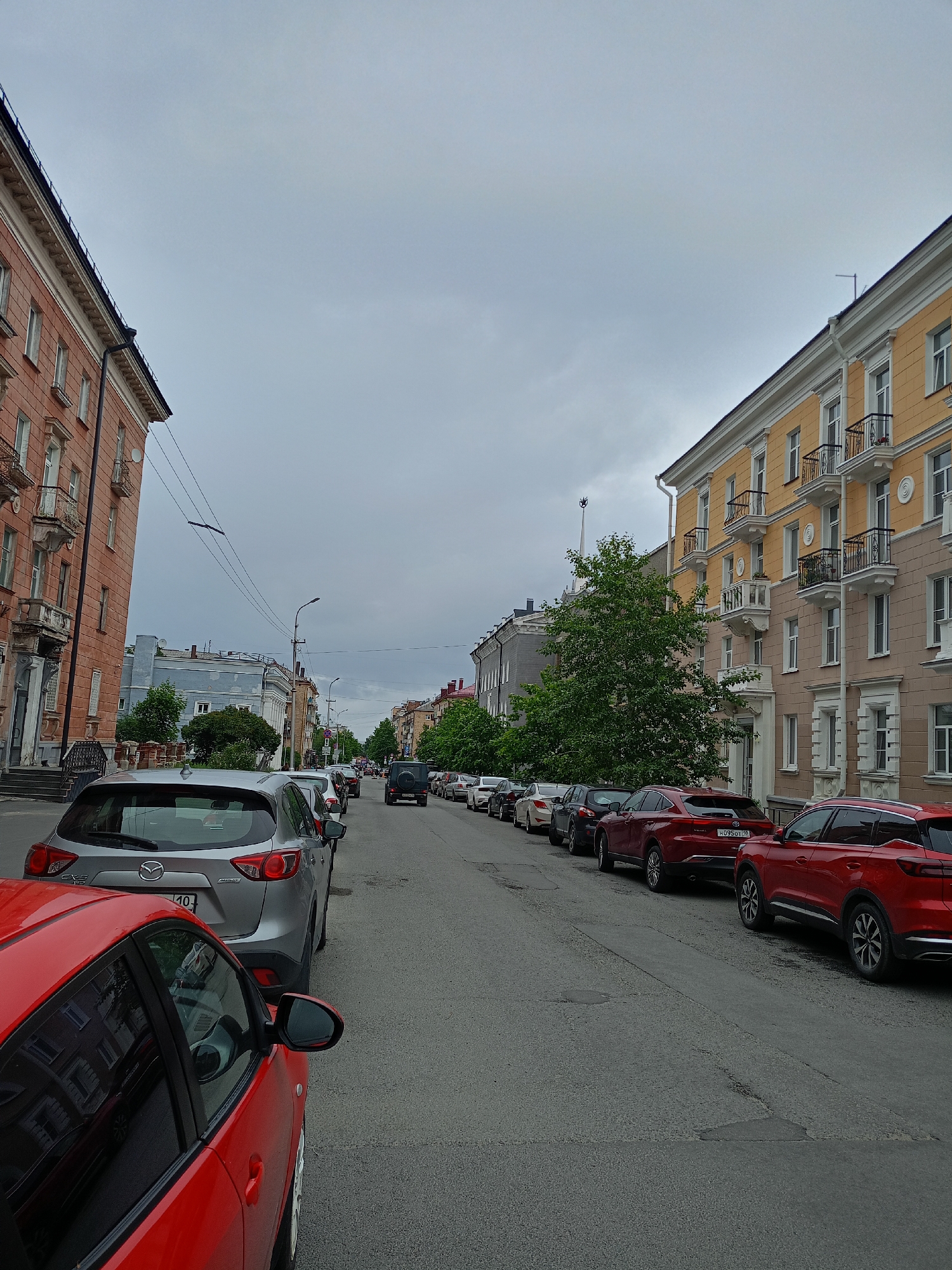The image shows a street scene in a city. The street is lined with buildings on both sides, most of which appear to be residential or commercial. Several cars are parked along the side of the street, and traffic appears to be moderate. The sky is overcast, suggesting a cloudy or rainy day. The buildings are of various architectural styles and colors, including a prominent pink building on the left and a yellow building on the right. Trees are visible along the street, and streetlights are present. The overall impression is a typical urban environment.