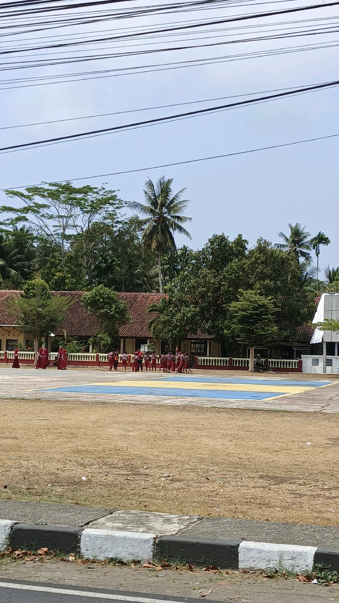 The image depicts an outdoor scene in Sukaresik, Indonesia, likely a schoolyard or community space. A group of children, appearing to be elementary school age, are gathered on a paved area marked with blue and yellow. Many of the children are wearing red uniforms, suggesting an organized activity or school event. In the background, there is a building with a terracotta-tiled roof, characteristic of Indonesian architecture, surrounded by lush green trees, including palm trees, indicative of a tropical climate. Power lines are visible crisscrossing the sky, a common sight in Indonesian urban and semi-urban environments. The ground in the foreground is dry and grassy, with a paved sidewalk and curb in the very bottom of the frame. The overall impression is of a bright, sunny day, with the children engaged in some form of activity in the schoolyard.