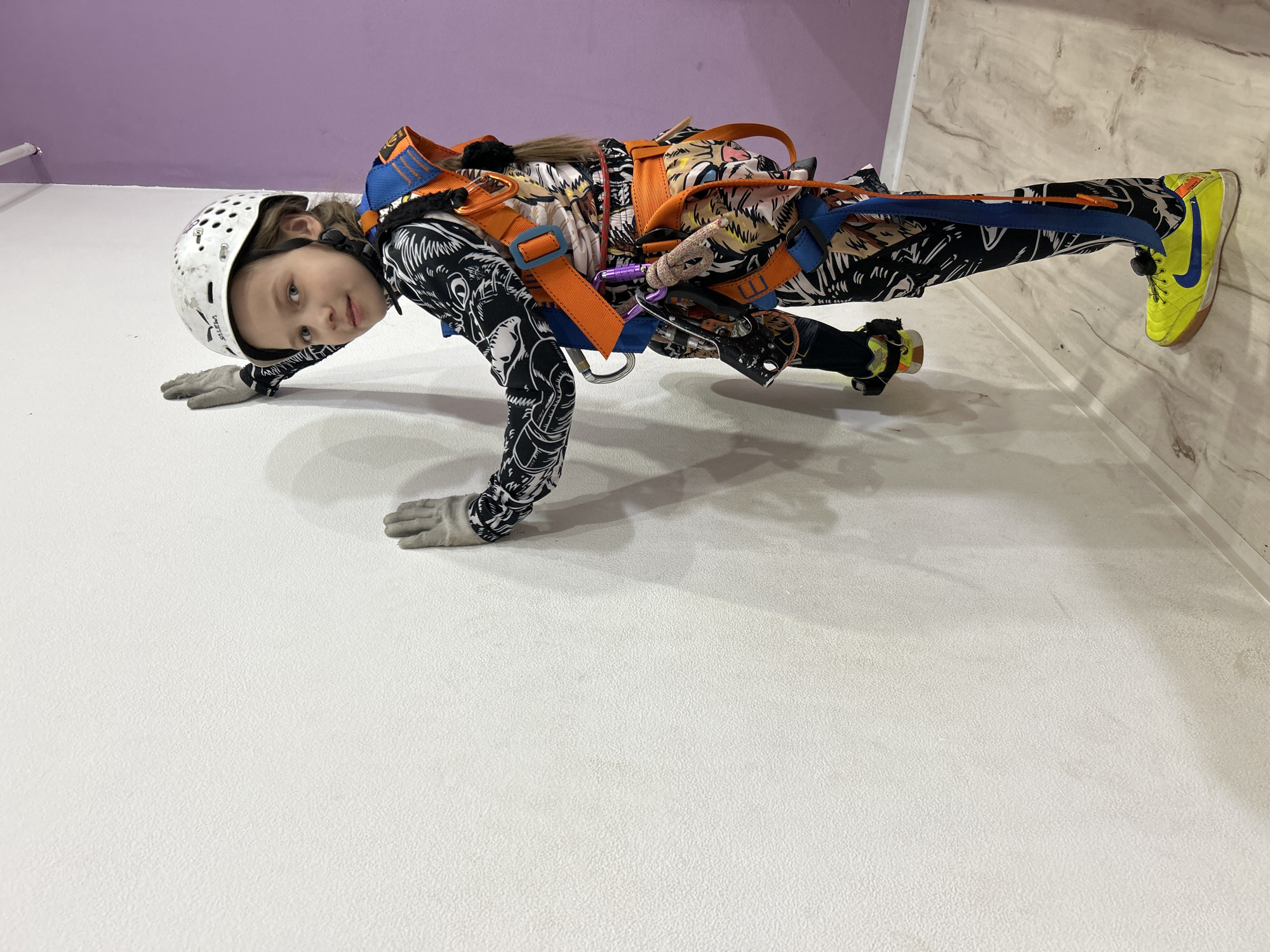 This media file captures a young child, likely a girl, engaged in an indoor climbing activity in Rusalevka, Russia. The image is oriented sideways, but if rotated, it shows the child in climbing gear positioned against a textured white climbing wall.
The child wears a white climbing helmet with "Salewa" visible on the side, indicating the brand. They are dressed in a long-sleeved, black and white patterned jumpsuit or separate top and pants, which features intricate designs. Over this outfit, an orange and blue climbing harness is securely fitted, equipped with various metal carabiners (including a purple one) and webbing loops. Gray gloves cover both hands, suggesting protection and grip for climbing. On their feet are bright neon yellow and blue athletic shoes, identifiable by the "Nike" swoosh logo on the side.
The child is seen leaning against the white textured climbing wall, with one hand and foot positioned as if actively climbing or in a resting stance. Their gaze is directed towards the camera, displaying a calm and composed expression. Above the white climbing wall, a smooth, light purple wall section is visible. To the right of the child's feet, the floor, made of a light-colored, patterned material resembling wood or laminate, can be seen. The overall setting is clearly an indoor climbing gym or adventure park, indicated by the specialized gear and the design of the wall. No specific time of day is discernible, but the indoor lighting suggests it is taking place during regular operating hours.