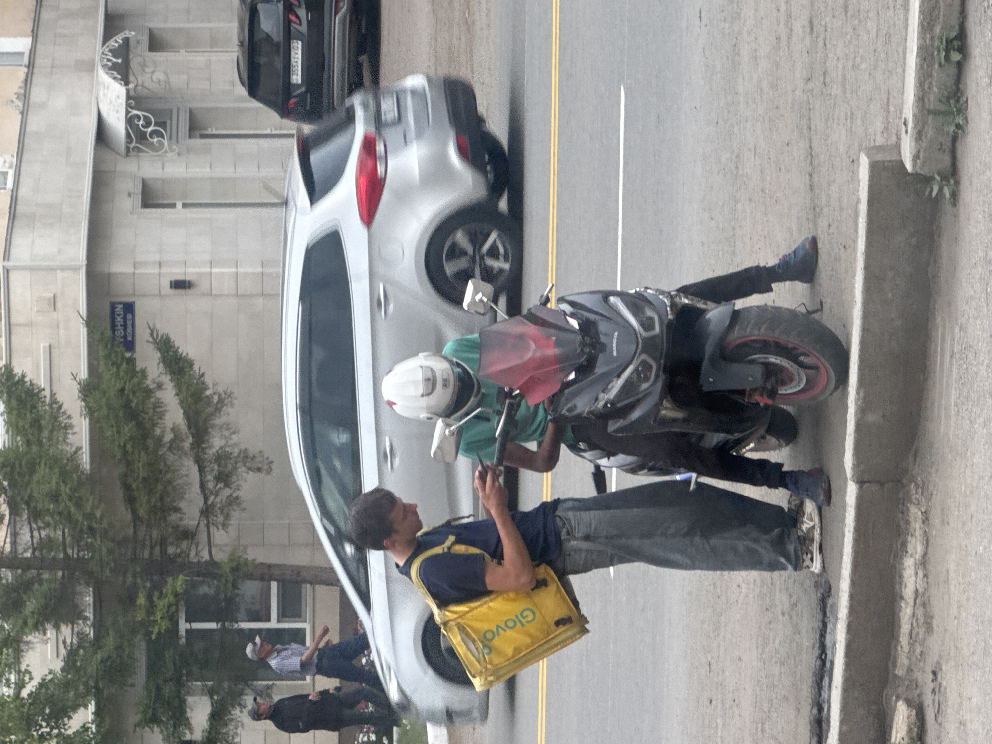 The image captures a street scene in Kokshetau, Kazakhstan. In the foreground, a young man wearing a dark blue t-shirt, light blue jeans, and a yellow "Glovo" delivery bag over his left shoulder is standing and looking at his smartphone. He is positioned next to a black scooter with a red windshield. On the scooter sits another individual wearing a green t-shirt and a white helmet. Behind them, a silver minivan is driving on the road, and a black SUV is parked on the right side of the image. In the background, a building with a sign reading "YSHKIN KOSHEE" is visible. Two older men, one in a dark jacket and the other in a plaid shirt and cap, are walking on the sidewalk to the left of the minivan. The environment appears to be an urban setting with a paved road and sidewalk.