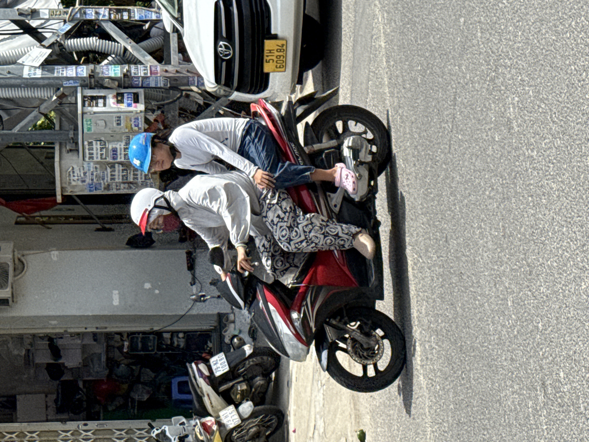 Two people are riding a red motorcycle on a paved road in Bac Nha Trang Ward, Vietnam. The person in front is wearing a white helmet with a red trim and a light-colored long-sleeved shirt, and is partially obscured by a patterned fabric. The person behind is wearing a blue helmet and a light-colored long-sleeved shirt, with blue pants and pink clogs. They are both looking forward. The motorcycle has the brand name "BLADE" visible on its side.
In the background, a white car with a license plate reading "51H 609.84" is parked. To the left of the motorcycle, another motorcycle with a license plate "79-N2 07362" is visible. The scene appears to be in an urban environment, with buildings and electrical poles in the background. The buildings have various signs and wires attached to them. The overall lighting suggests it is daytime.