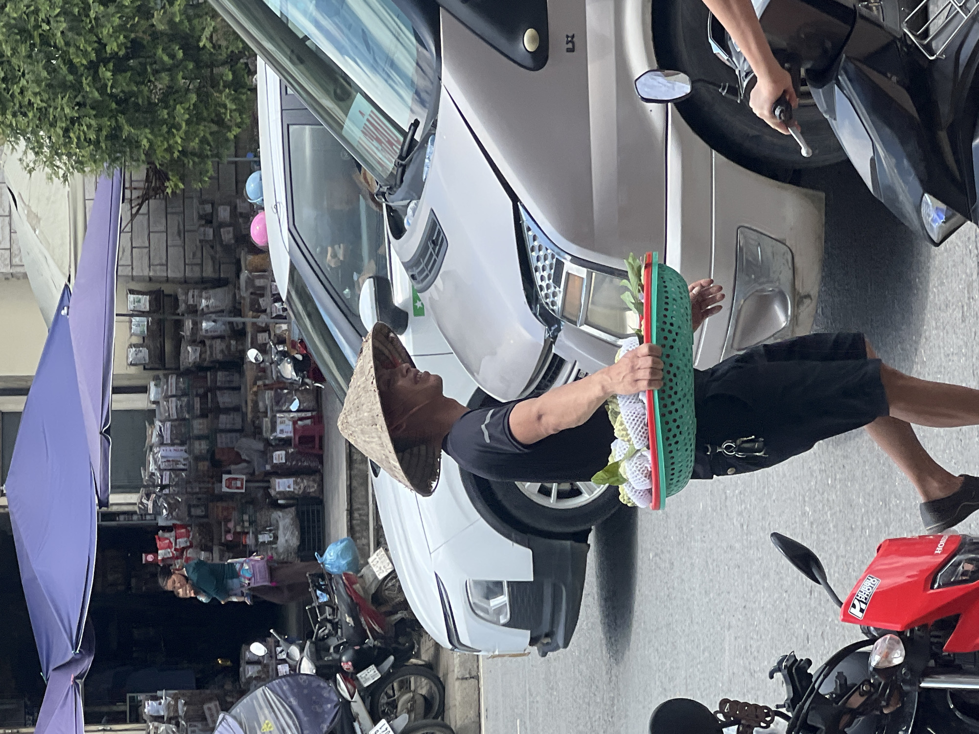 A man wearing a conical hat and a black t-shirt walks on a street in Sa Pa Ward, Vietnam, carrying a green basket filled with wrapped fruit. He is walking past a white car and a beige van. In the background, there are shops with awnings and displays of goods. Several motorbikes are parked on the side of the road, and a woman is seen holding a baby in front of one of the shops.