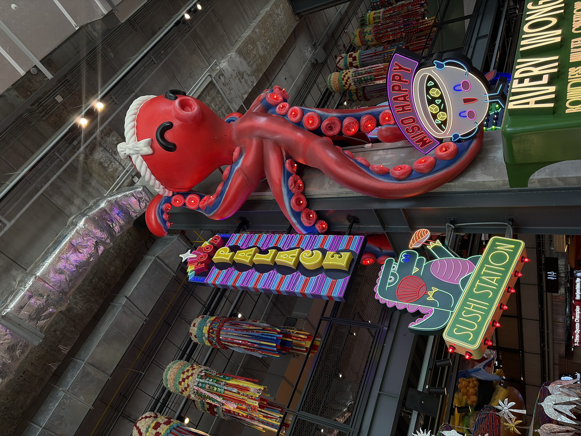 The image showcases an eclectic display of illuminated signs and decorations hanging from the ceiling within a marketplace setting. Prominently featured is a large red octopus figure adorned with blue accents and illuminated red circles along its tentacles. Adjacent to the octopus is a sign that reads "MISO HAPPY" above a cartoonish bowl of soup. A green rectangular sign states "AVERY WONG," followed by the text "ROUNDLESS MULTED CREMONE" in smaller print below.
Further below, a yellow neon sign spells out "FOOD PALACE" on a blue and pink striped background. To the right, another sign reads "SUSHI STATION" in green neon, accompanied by a whimsical illustration of a dinosaur holding a piece of sushi.
Hanging beneath the signs are several colorful, cylindrical decorations made of patterned fabric. The overall style of the scene is vibrant and playful, with a mix of food-related signage and artistic elements.
Based on the signs and decor, this image most likely originates from ICONSIAM in Bangkok, Thailand. The signs for "Sushi Station", "Food Palace", and "Avery Wong" are restaurant and establishment names within the complex.