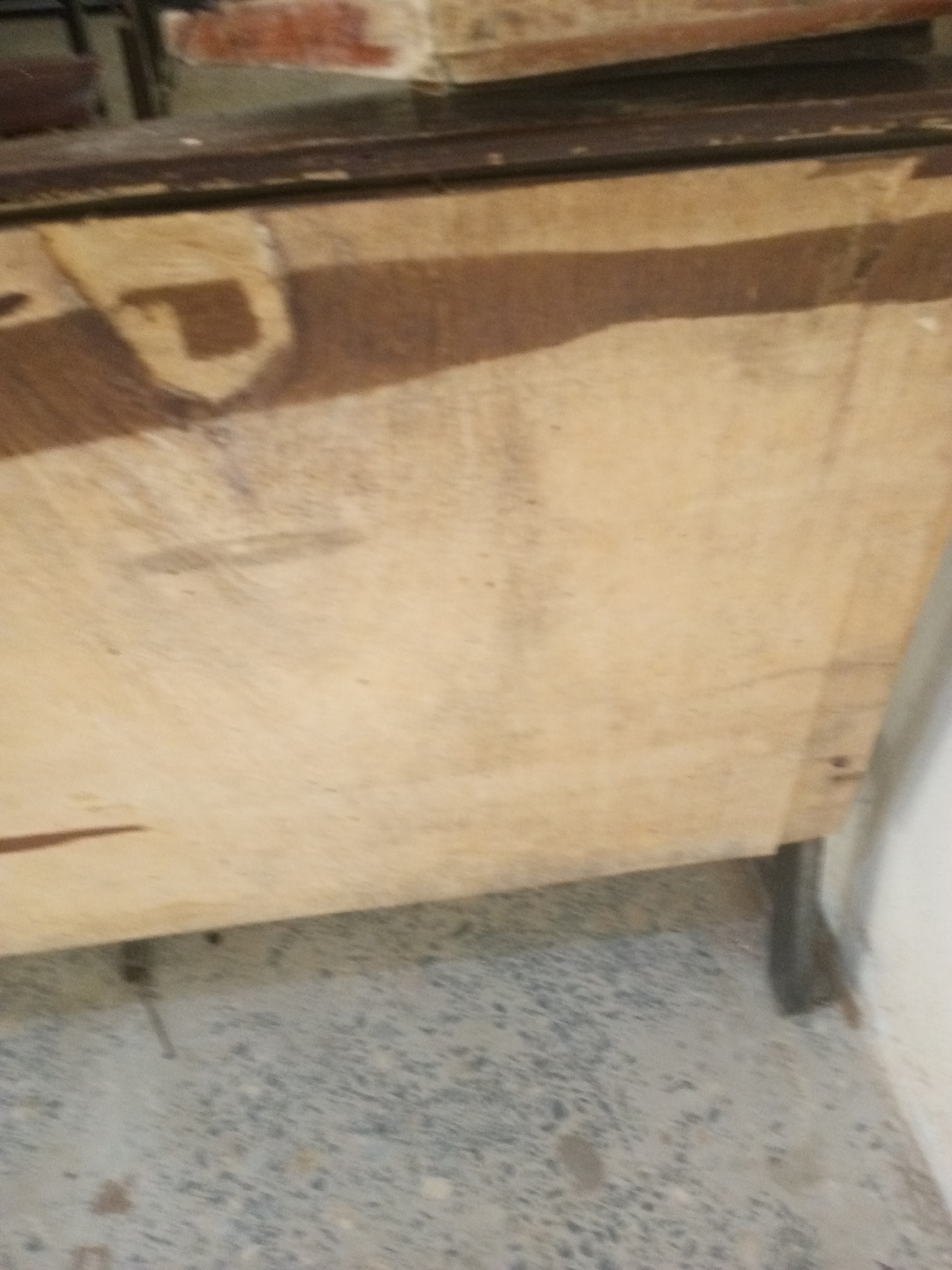 A close-up, low-angle shot shows the side of a wooden chest or cabinet. The wood grain is visible, with lighter and darker shades of brown. The chest has a dark-colored metal edge along its top. In the upper left of the image, a portion of a brick or stone wall is visible, with some red and white coloration. The chest rests on a dark metal support leg on the right side, and on the floor, which appears to be a rough, textured surface like concrete or gravel. The scene is indoors, with a bright light source casting shadows on the chest. The image is unfocused and grainy, making it difficult to discern fine details. The location is identified as Jalingo, Nigeria.