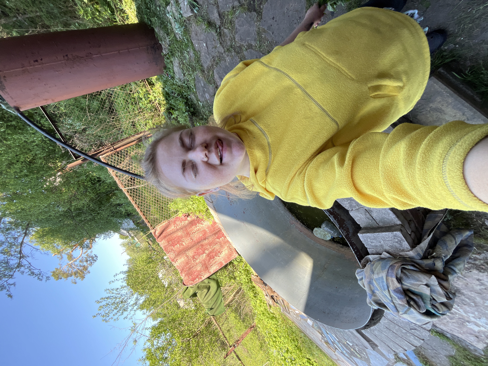 A woman in a yellow fleece jacket is lying down, taking a selfie. Her eyes are mostly closed, and her tongue is sticking out. The location appears to be outdoors, possibly in a garden or yard. Visible elements include a metal container (possibly a barrel), a fence, greenery (trees and shrubs), paving stones, a boat, and some other objects covered in fabric. The scene is sunny. Without specific landmarks or location metadata in the image, pinpointing the precise location in Chaikovsky, Russia, is impossible.