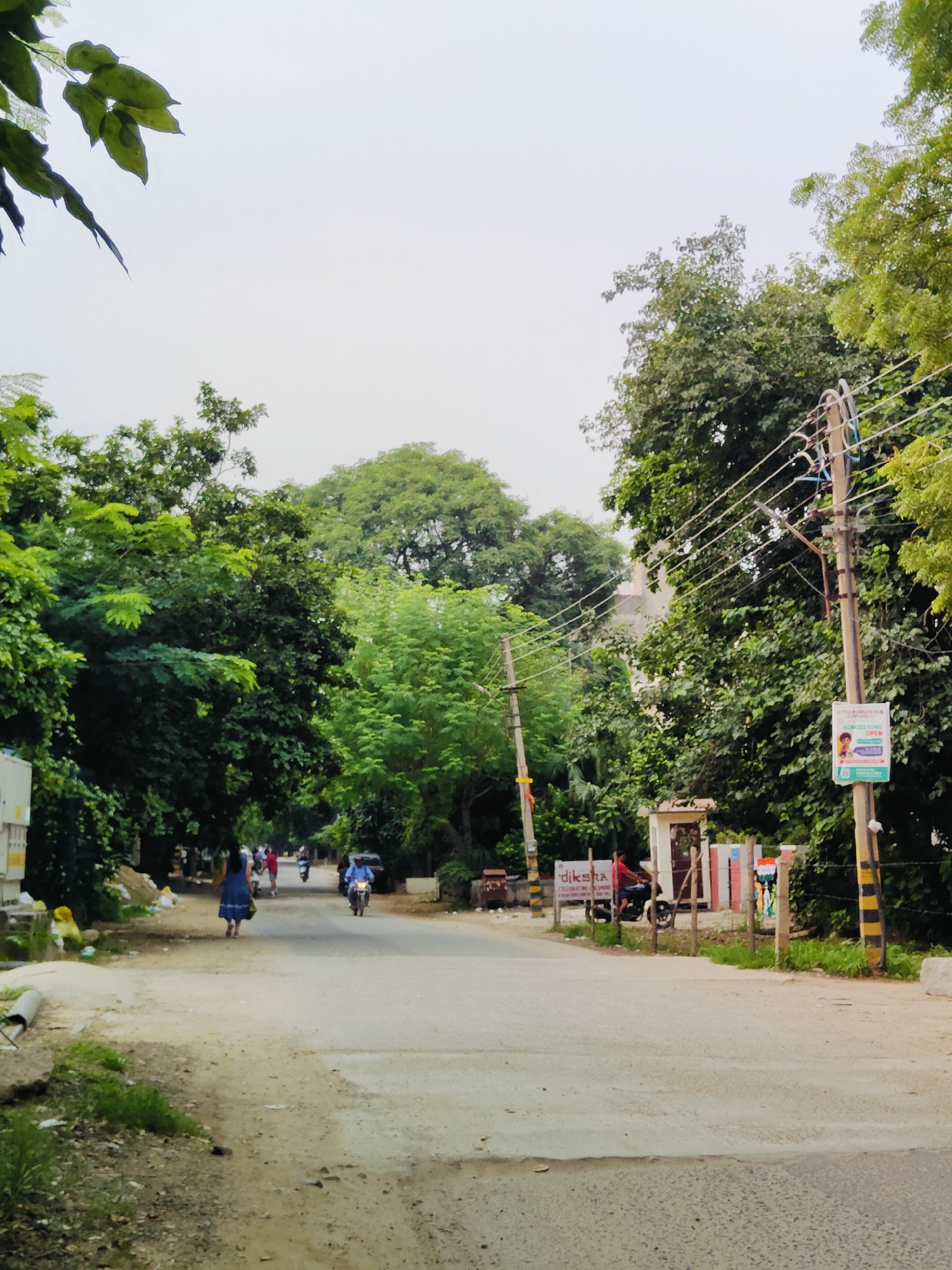 The image depicts a moderately populated street scene in Gurgaon, India, on an overcast day. The road is paved but shows signs of wear and tear, with patches of dirt and debris. Lush green trees line both sides of the street, with a prominent power pole featuring overhead wires and a sign on the right side.
In the foreground, the road stretches into the distance, with a few vehicles and pedestrians visible. On the left, a woman in a blue dress walks away from the camera, carrying a bag. Further down the road, several people are seen on motorcycles and bicycles, moving in the same direction.
To the right, a small booth-like structure stands next to the road, with a sign reading "DIKSHA" and another poster with "ADMISSIONS OPEN" in English. A person in a red shirt is seated on a motorcycle in front of this structure. The background reveals more dense foliage and buildings, indicating an urban or semi-urban environment. The overall atmosphere is one of everyday activity on a somewhat unkempt street.