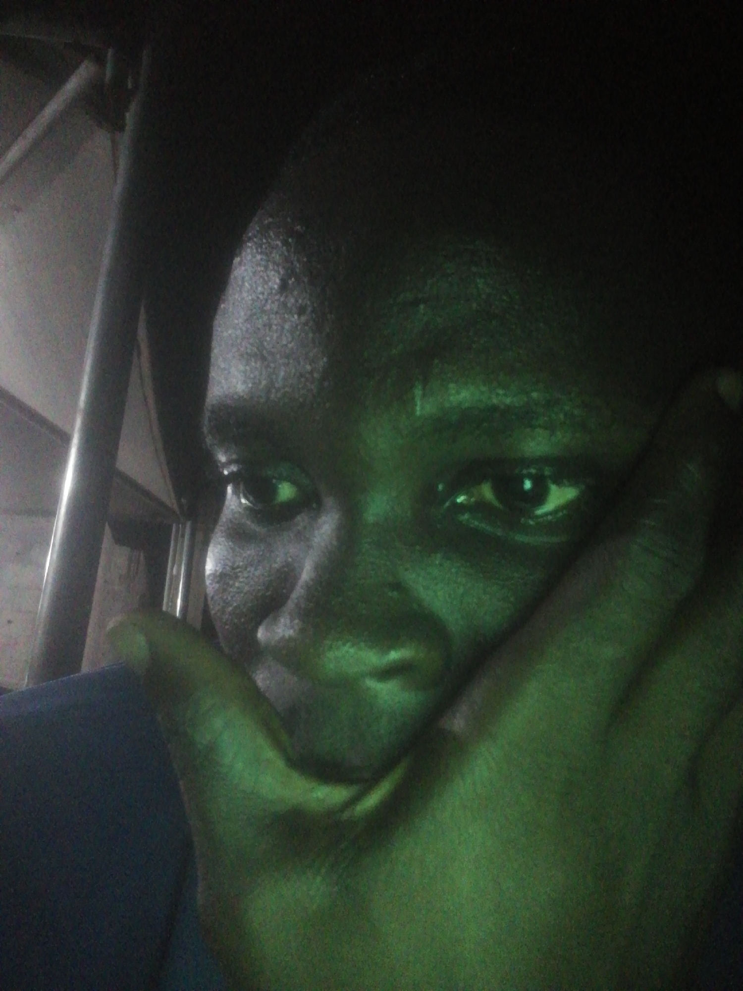 A close-up selfie of a dark-skinned person with their face partially obscured by their right hand. The person's eyes are visible and appear to be looking slightly off-camera. The lighting has a green tint, making details difficult to discern. A metal bar is visible in the background on the left. The specific location in Ikorodu, Nigeria, cannot be determined from the image.