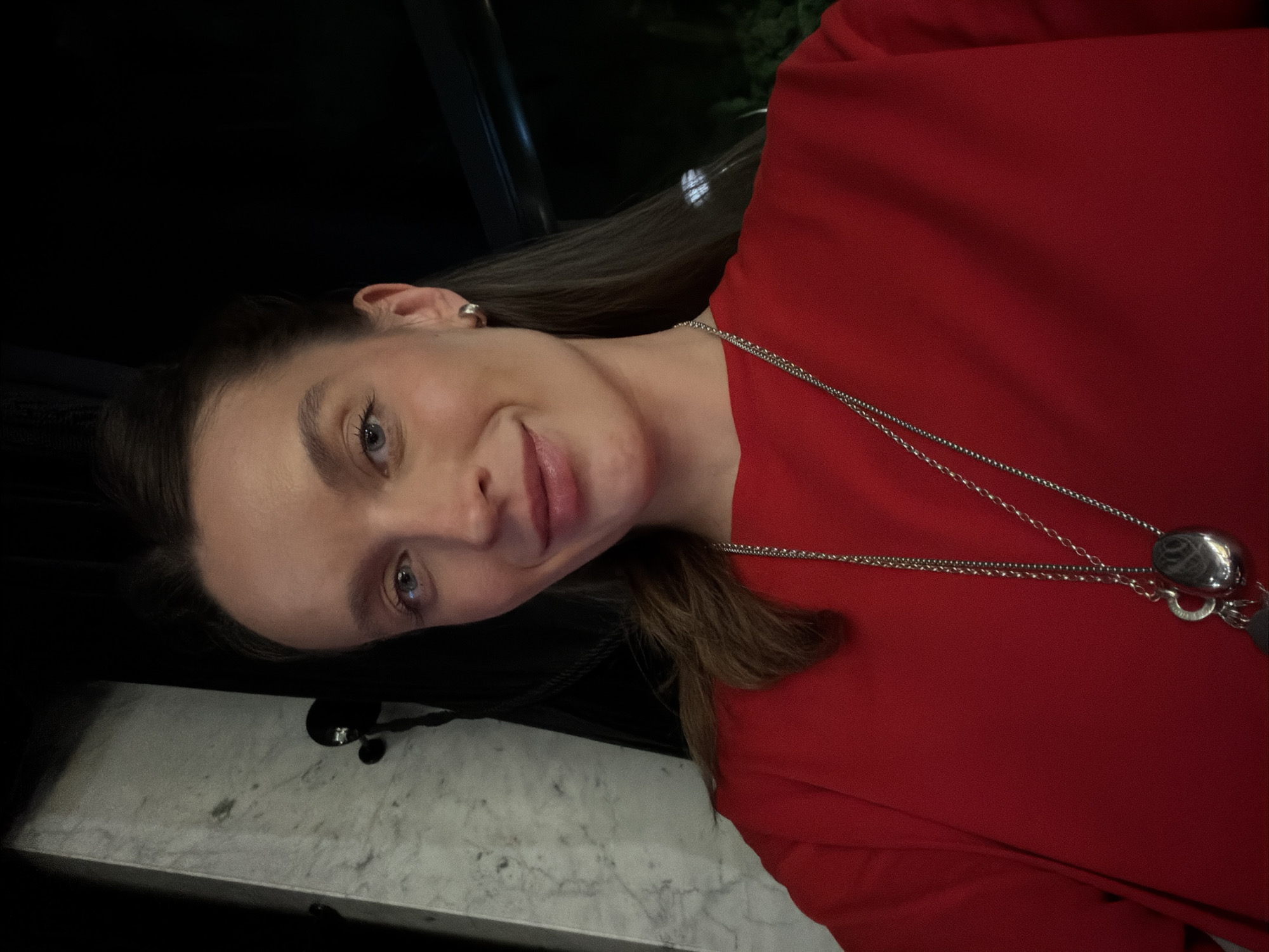 A woman with light skin and blonde hair is shown in a close-up portrait. She is wearing a red shirt and a silver necklace with a pendant. The background is dark and appears to be indoors, with a marble table surface visible in the lower left corner. The location and context of the image within the city of Budva, Montenegro, cannot be determined based on the visual information provided.