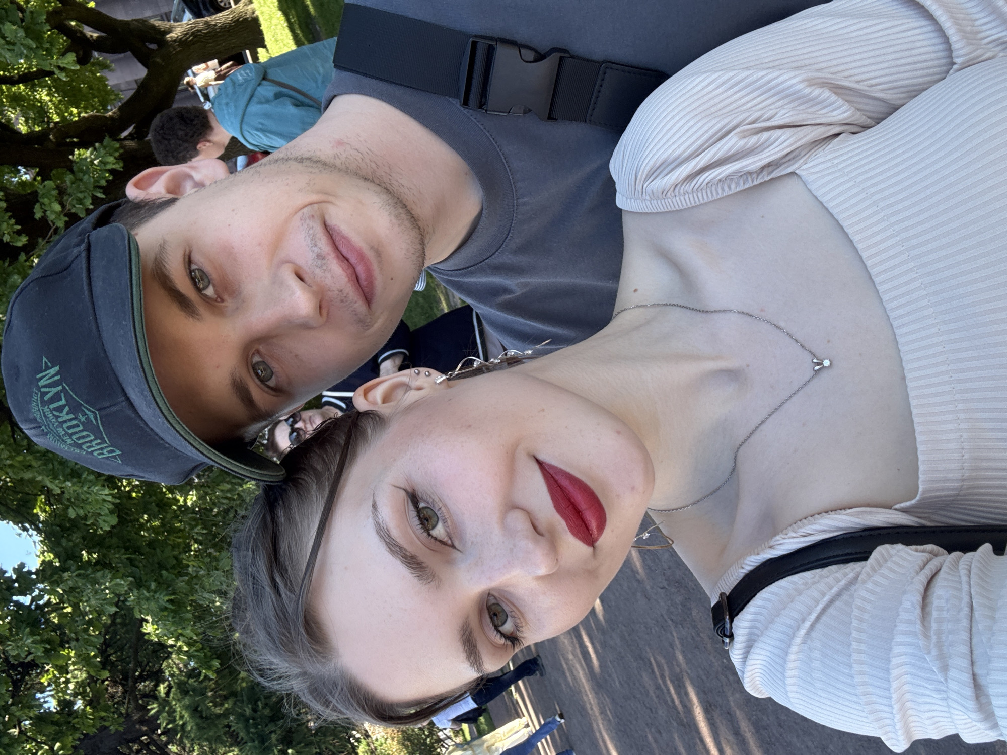 This outdoor photograph, taken in a public green space within Saint Petersburg, Russia, captures two young adults in a selfie-style composition on a bright, sunny day. In the foreground, a female with fair skin, dark hair pulled back, green eyes with dark winged eyeliner, and bright red lipstick smiles towards the camera. Her visible left ear features multiple piercings and a long, spiraling silver-toned earring. She wears a light beige or pink ribbed square-neck top, a delicate silver chain with a bunny pendant, and a black strap from a bag across her right shoulder. Behind her, a male with fair skin, light stubble, and green-grey eyes looks towards the camera with a slight smile. He wears a dark grey t-shirt and a dark baseball cap with "BROOKLYN" in green lettering, with a black backpack strap across his left shoulder featuring a plastic buckle. The background consists of lush green deciduous trees, a paved pathway, and several other individuals, including a partially visible woman in sunglasses and another person in a light blue hooded jacket seen from behind. Strong sunlight illuminates the scene, casting clear shadows.