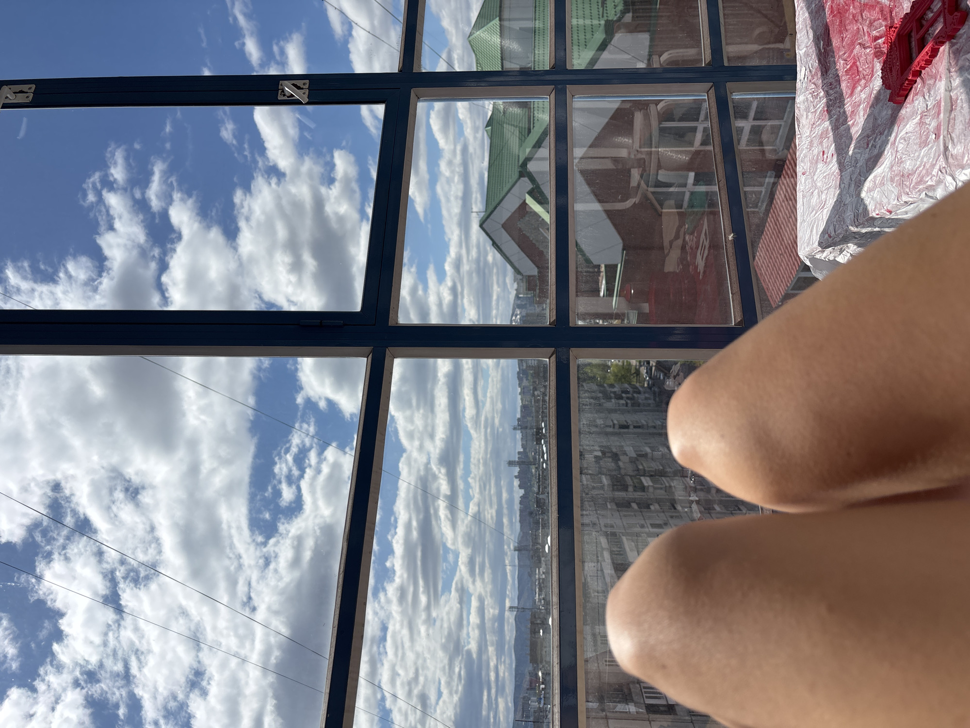 The image shows a view from a high vantage point, possibly inside a building with large windows, overlooking a cityscape.
**Environment:**
* **Setting:** Likely an urban area, discernible from the presence of multiple buildings.
* **Sky:** Visible through the windows, featuring a sky with numerous clouds.
* **Windows:** Grid-like windows with dark frames.
**Objects:**
* **Building Reflections:** Reflected in the windows are buildings with different architectural styles, one with a green roof.
* **Cityscape:** Distant view of the city with multiple buildings, possibly Krasnoyarsk, Russia.
* **Red-painted object:** Upper right side has a red-painted object on top of the white plastic surface.
**People/Subjects:**
* **Person:** Two knees of a person are in the shot in the lower right corner.