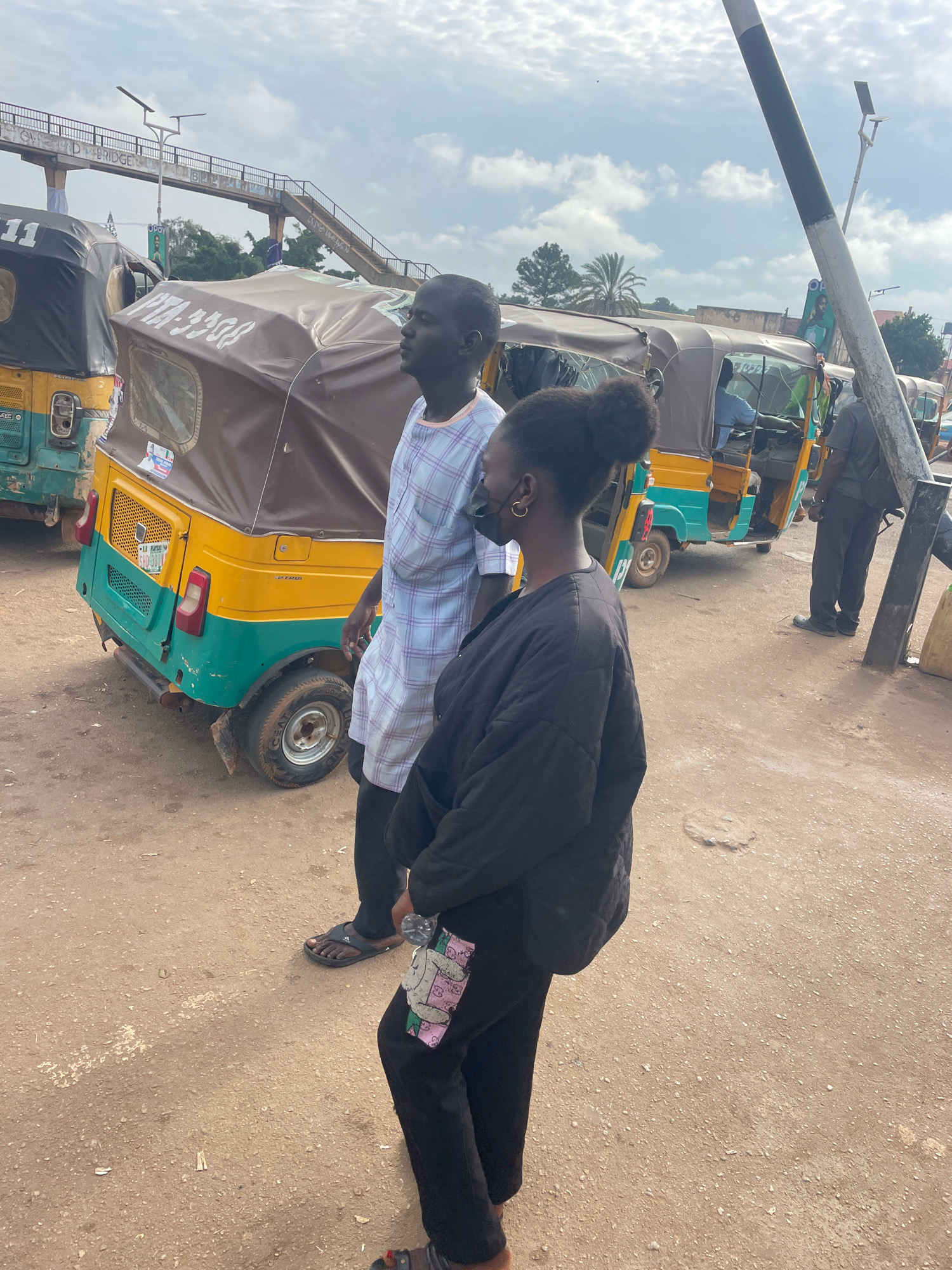 In Gura Top, Nigeria, two individuals, a man and a woman, stand near a row of auto-rickshaws. The man, wearing a blue and white checkered shirt, faces to the right, with his profile visible. The woman, in a black jacket and pants, stands closer to the camera, looking to her right. Her dark hair is pulled up in a bun, and she wears a black face mask and gold hoop earrings.
Several auto-rickshaws, commonly known as "keke napep" in Nigeria, are parked in a dirt lot. One auto-rickshaw in the foreground is predominantly yellow and green, with a brown canopy. Its license plate reads "SHD-3614". Other auto-rickshaws are partially visible behind it, some with drivers inside.
In the background, a pedestrian bridge spans overhead, with the word "BRIDGE" visible on it. The sky is overcast with scattered clouds, and palm trees and other vegetation are visible in the distance. A tall pole with black and white diagonal markings stands to the right of the frame, near the auto-rickshaws. The ground is a dusty, reddish-brown dirt.