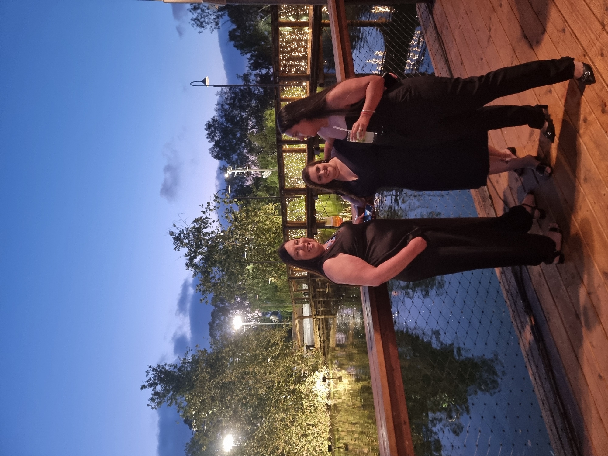 Three women are standing on a wooden deck at dusk, by a body of water. The woman on the left is wearing a black jumpsuit and is holding a glass. The woman in the middle is wearing a black dress and has her arm around the woman on the right. The woman on the right is wearing a black jumpsuit and is holding a drink with a straw. In the background, there is a bridge decorated with string lights, trees, and streetlights. The setting appears to be a restaurant or event space in Ga'ash, Israel.