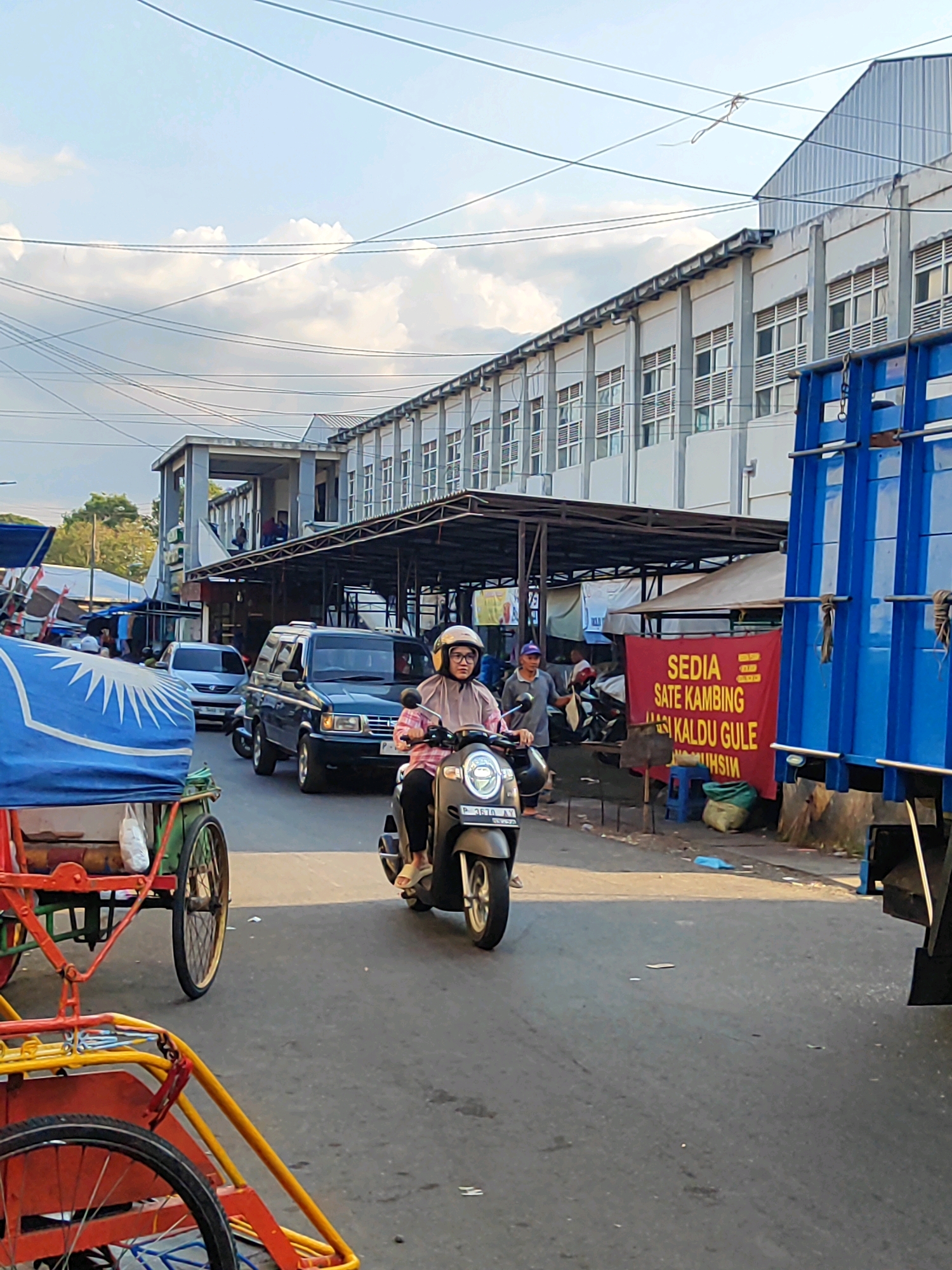 A busy street scene in Bondowoso, Indonesia features a woman riding a scooter. She is wearing a pink shirt, black pants, a light brown headscarf, and a white helmet. She is looking forward and smiling. The scooter is a dark gray Honda. In the foreground, a brightly colored pedicab with a blue tarp is visible. Behind the scooter, a dark blue truck is parked on the right side of the road, its side displaying a red banner advertising "SEDIA SATE KAMBING NASI KALDU GULE" which translates to "READY GOAT SATAY GOULASH BROTH RICE". A dark colored SUV is also present on the road, as well as other vehicles and market stalls. The background shows a multi-story building with a light-colored facade and numerous windows, likely a market or commercial establishment. The sky is partly cloudy with clear blue patches. Electrical wires crisscross the sky above the street.