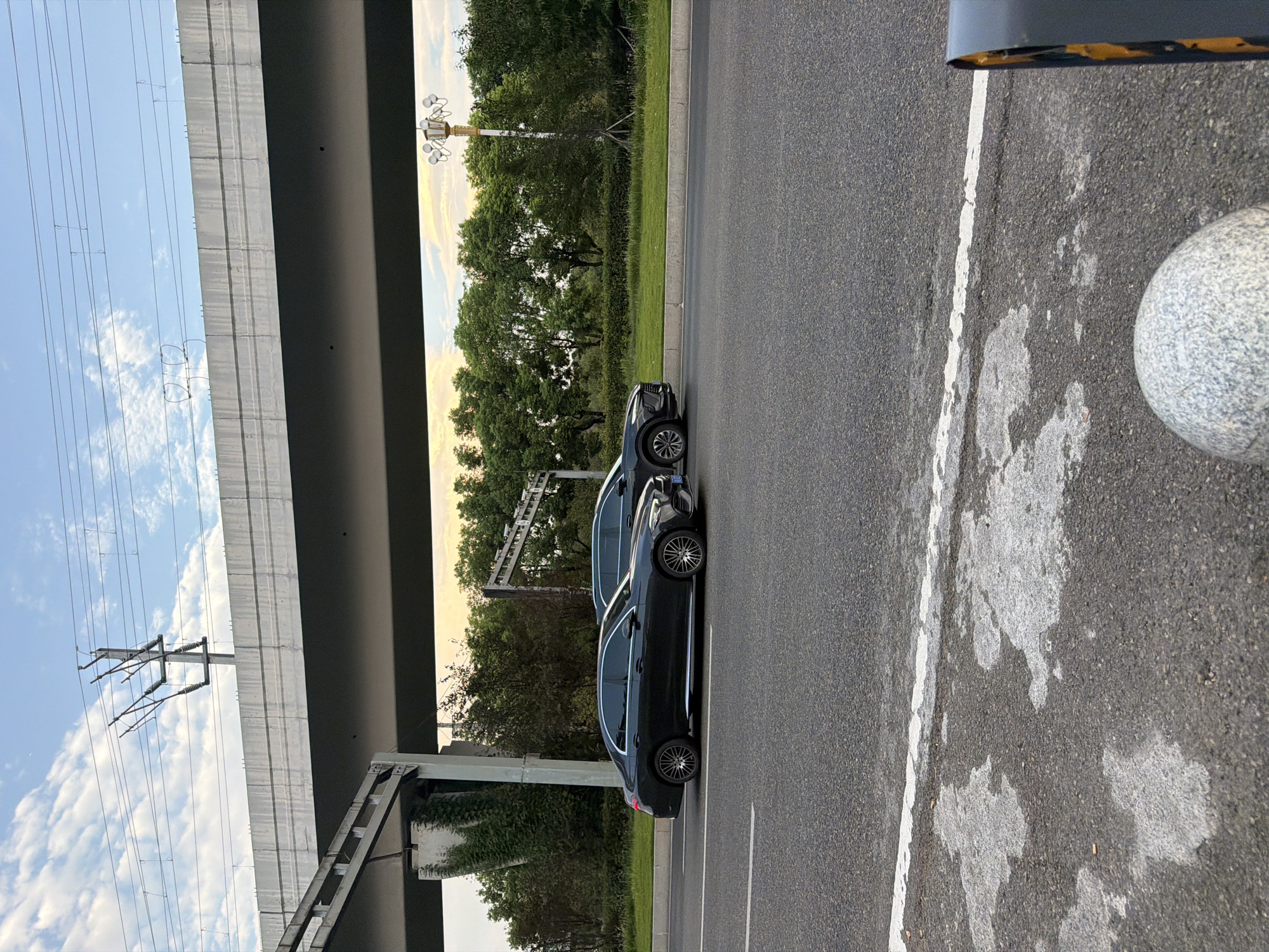 The image shows two cars, a black sedan and a dark SUV, driving on a road in Jiangjin, China. The cars are driving under an elevated structure, possibly a highway or railway overpass. There are trees and vegetation lining the side of the road. The sky is cloudy with the sun setting. The road is asphalt and has a white line marking the lane. In the foreground, there is a white patch on the road and a bollard.