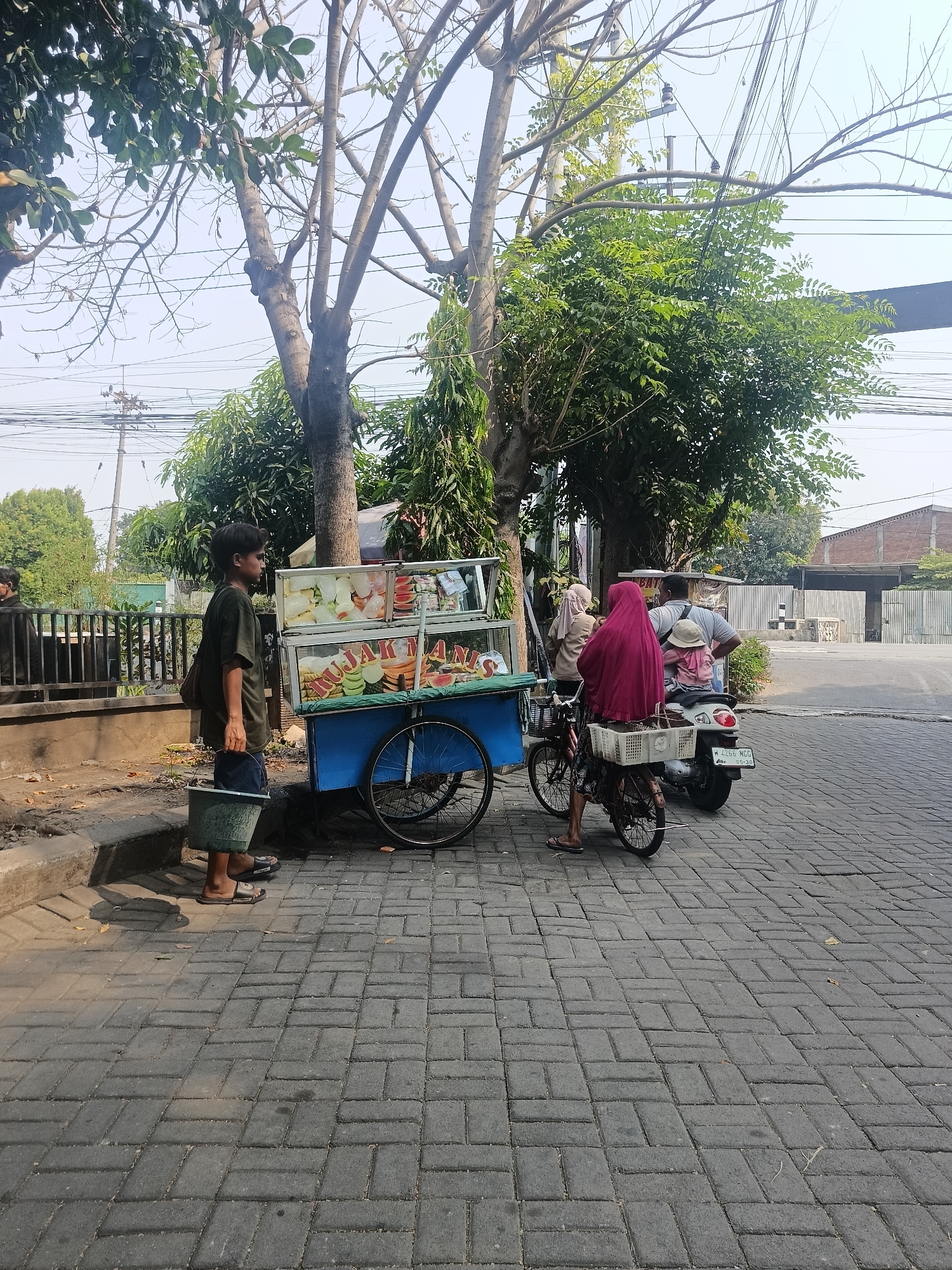 This image captures a street scene in Wedi, Indonesia, on a sunny day. A young boy stands to the left of a blue food cart, holding a green bucket. The cart is filled with various fruits, including watermelon, cantaloupe, and what appear to be ice-filled bowls, suggesting it is a fruit stand. The cart has a sign with Indonesian writing. To the right of the cart, a woman wearing a red hijab is seated on a bicycle, and another person, partially visible, is behind her. A white basket is attached to the back of the bicycle. Behind the bicycle, a scooter is parked. The ground is paved with interlocking brick pavers. Trees with green foliage are visible in the background, and there are power lines overhead. A building with a brick facade and a metal gate is visible in the distance, along with a fence. The overall atmosphere is that of a typical street in a small Indonesian town.