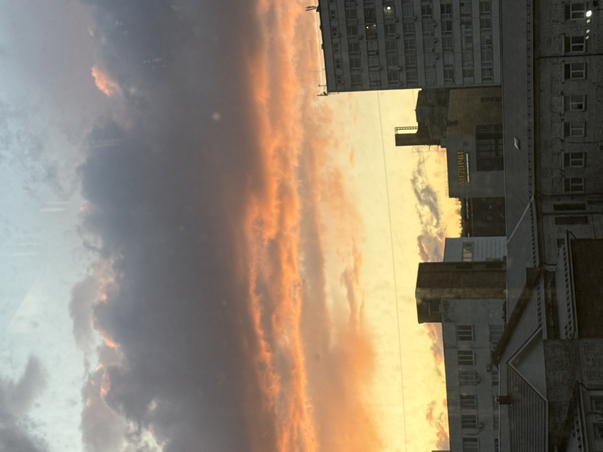 The image shows an urban landscape at sunset, likely in Krasnoyarsk, Russia. The right side of the image features several tall buildings with a mix of architectural styles and materials, including concrete and brick. A building with the Russian word "МЕДИЦИ" (likely part of "Медицина," meaning medicine) is visible. The left side of the image is dominated by clouds tinged with orange and yellow hues from the setting sun, contrasting with the grey and blue sky. There are no people visible. The focus is on the architectural elements and the sky's atmospheric conditions.