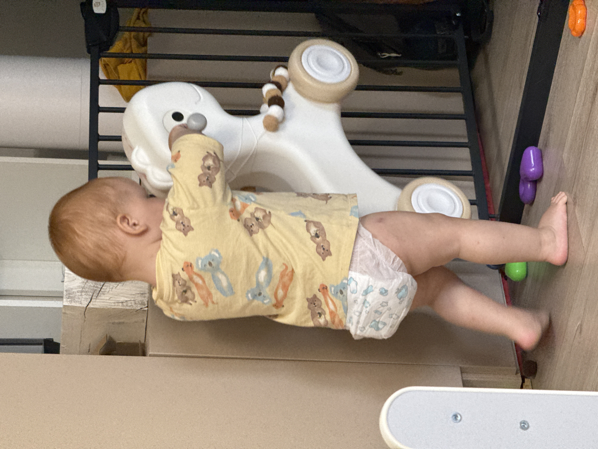 The image captures an infant, viewed from the back, with light reddish-brown hair. The infant is wearing a long-sleeved yellow shirt adorned with cartoon animal prints, including brown, orange, and light blue figures resembling koalas and foxes. A white disposable diaper featuring light blue patterns is visible. The infant is barefoot, standing on a light brown, wood-patterned floor.
The infant is actively engaging with a white, stylized plastic toy, potentially a push or ride-on toy, which bears resemblance to an animal with a rounded head, a dark eye, and a prominent grey nose or button. This toy is equipped with light wooden-colored wheels that have white central hubs. A decorative, segmented coil in shades of brown, white, and dark brown is attached to the toy's side. The infant's hands are placed on the upper part of the toy, suggesting either support or manipulation. The slight blur evident in the infant's right foot indicates motion.
In the background to the left, a vertical light beige wall or furniture panel is visible. Adjacent to this, a vertical architectural element with a rough, textured, and cracked surface, possibly a weathered wooden beam, can be seen. To the right, a black metal playpen or safety gate with vertical bars extends across the scene. Behind this gate, a piece of crumpled yellow fabric and a dark, amorphous object are discernible.
Various small plastic toys are scattered on the floor: a bright green spherical object near the infant's left foot, two purple oval-shaped objects positioned near the base of the black gate, and an orange star-shaped or flower-shaped object further to the right.
No visual information is present within the image to identify its geographic location as the city of Izhevsk, Russia, or any other specific region or country.