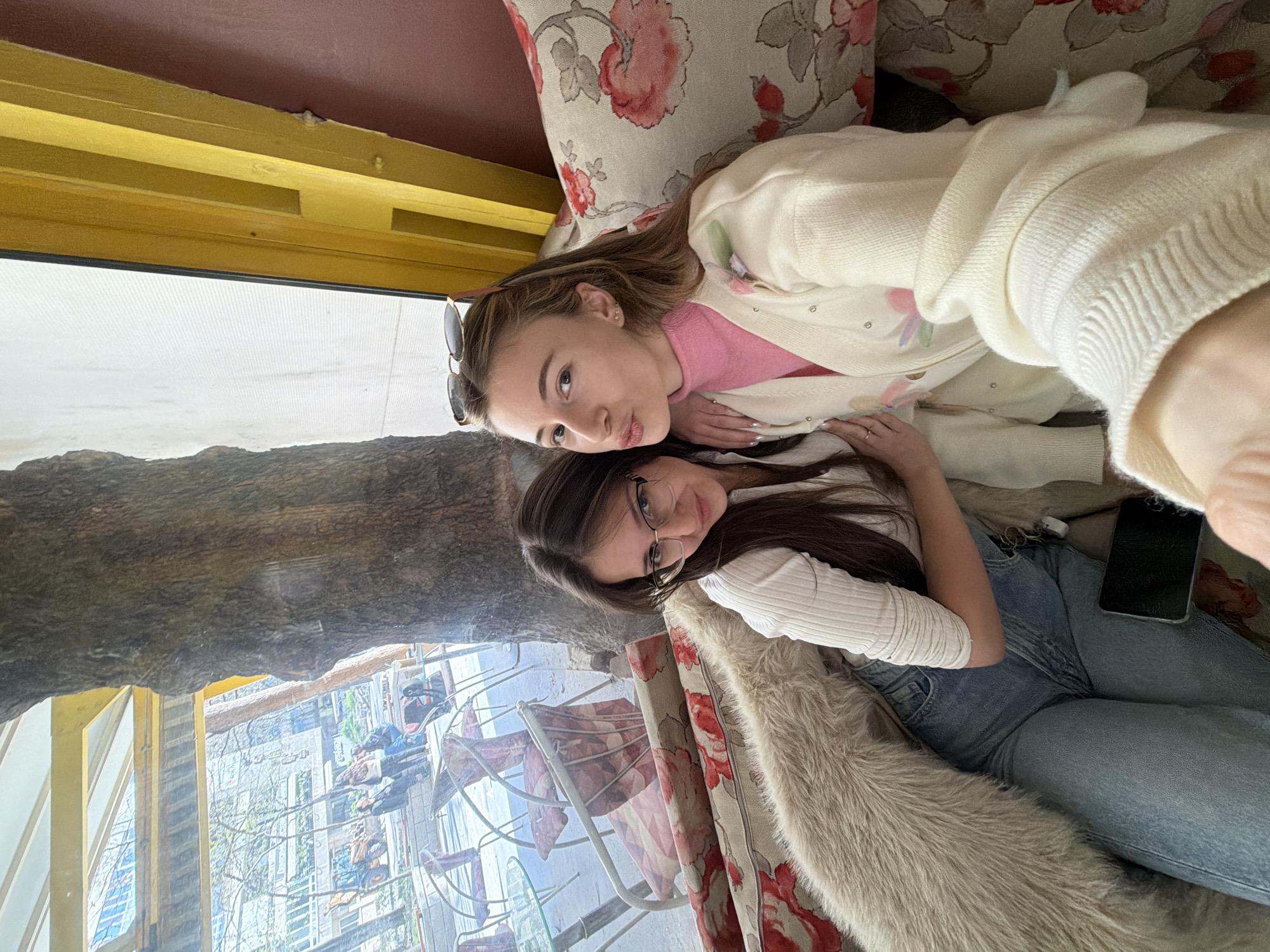 Two young women pose closely together in an interior setting. The woman on the left wears a pink shirt, a white cardigan, and sunglasses perched on her head. Her hair is light brown. The woman on the right wears a white long-sleeved shirt, glasses, and blue jeans. Her hair is dark brown. They are both lying on furniture adorned with floral patterns and faux fur. A black smartphone lies near the woman in blue jeans.
Through a nearby window, a street scene is visible. Buildings and trees line the street, and people are visible walking and seated at outdoor tables. The setting is identifiable as located in Plovdiv, Bulgaria, due to the architecture and urban layout visible in the window.