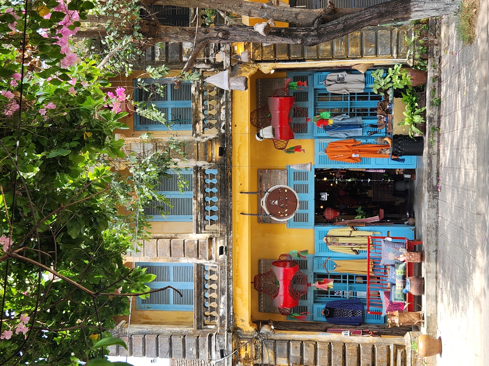 This is a frontal medium shot of a shop exterior in Hoi An Ancient Town, Vietnam. The building has a weathered yellow facade with ornate architectural details. Blue shutters frame the shop's windows and doorway. Green foliage with pink blossoms hangs from overhead branches, casting shadows on the scene.
The shop displays clothing items on hangers in the windows and doorway, including a dark blue jacket with a pattern, a yellow dress, a yellow kimono-style garment, an orange robe, a grey jacket, and a light grey shirt. A red bench with several colorful pillows is positioned in front of the central doorway. Baskets and decorative items are placed on and around the bench.
Further inside the shop, lanterns and various other goods are visible. A large, abstract red art installation hangs above the doorway, accompanied by several smaller decorative elements. A sign with the word "HART" is suspended from the facade. A tree with rough bark stands to the right of the shop. The ground in front of the shop is paved with square tiles, and a few fallen petals are scattered on the pavement. There are no people visible in the image.