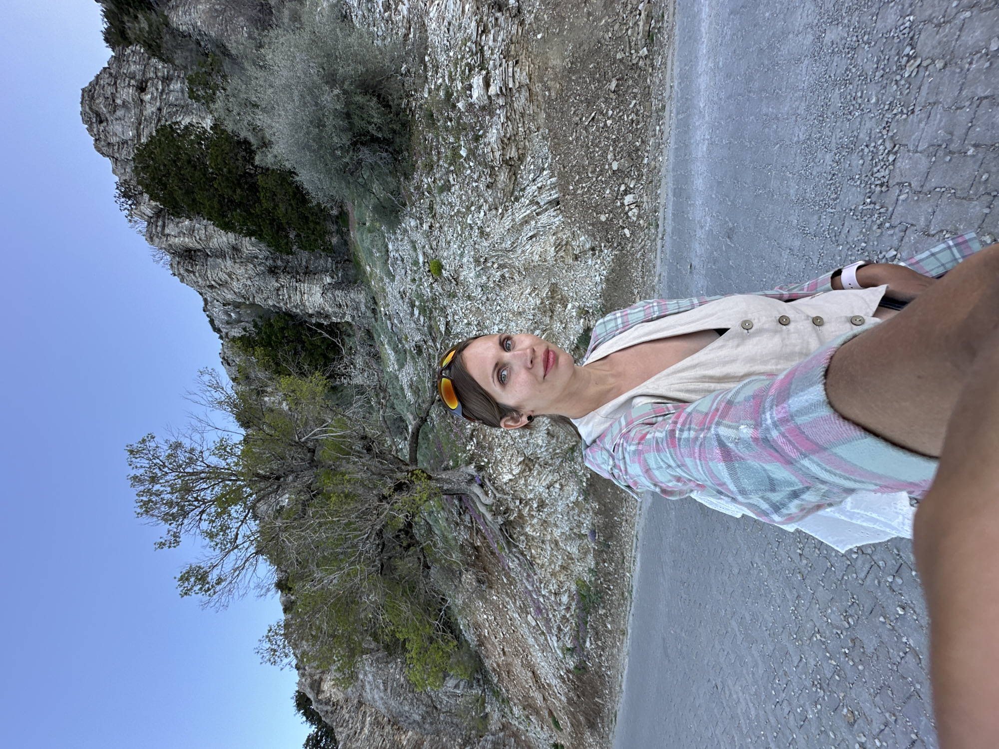 A woman in a plaid shirt and khaki vest stands on a concrete walkway beside a cliff in Fethiye, Turkey. The cliff is composed of layered gray rock and sparse vegetation. A leafless tree clings to the cliff face. The sky is visible in the background. The concrete walkway shows signs of wear and scattered debris.