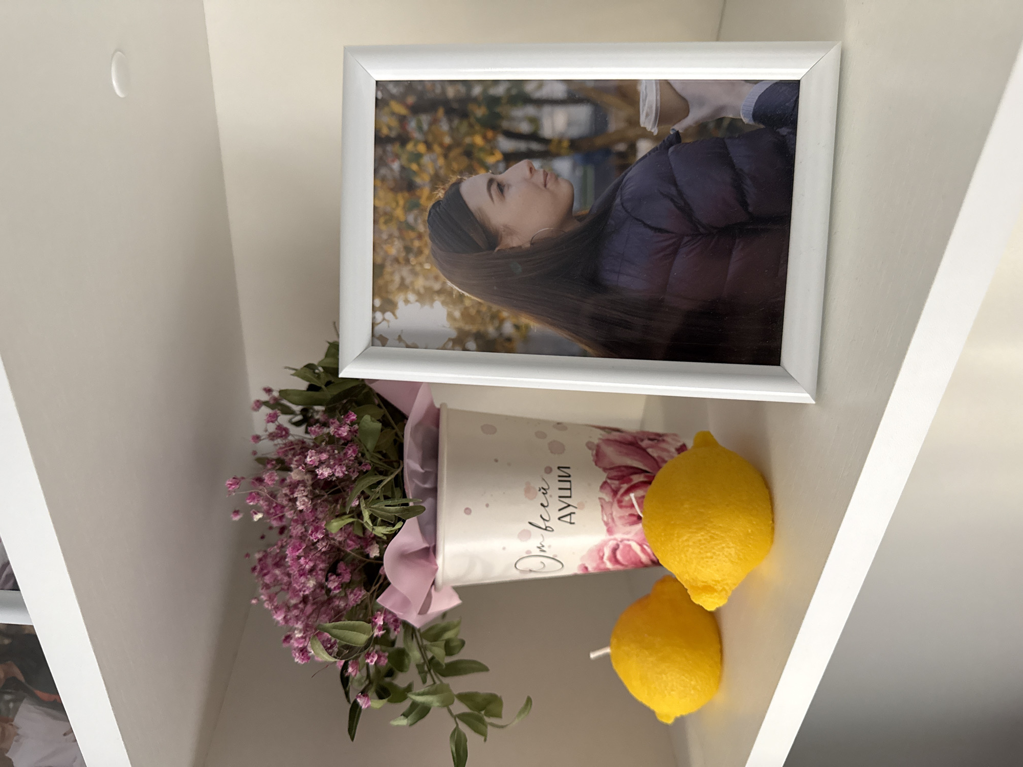 A white shelf contains a framed photograph, a bouquet of pink flowers in a white vase, and two yellow lemons. The photograph is of a woman in a puffer jacket, taken outdoors amidst trees with autumn foliage. The text on the vase is in Russian. The background is a plain white wall. While there is no explicit evidence of the location being Moscow, Russia, it cannot be definitively ruled out.